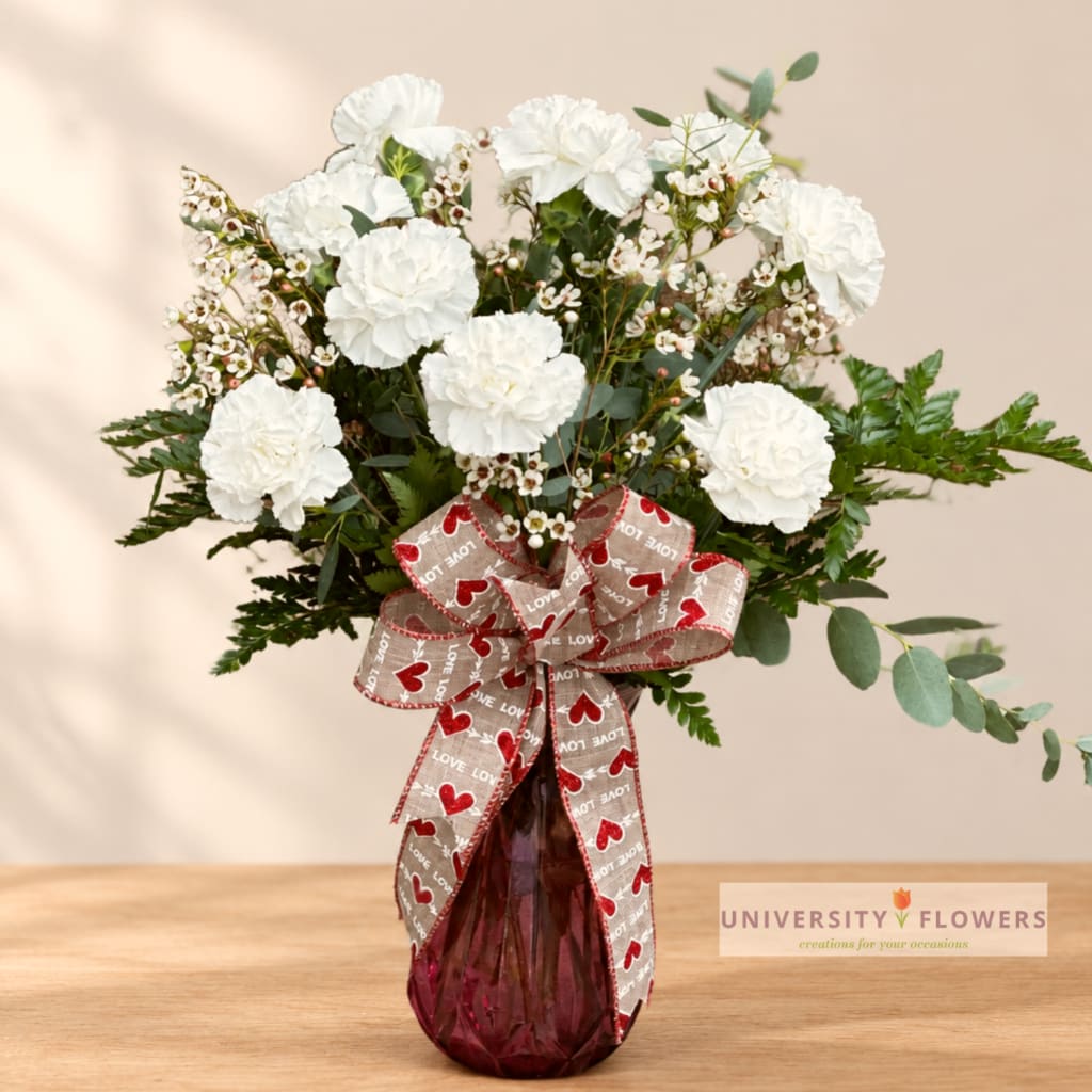 White carnations and filler flowers in a burgundy glass vase with a heart ribbon