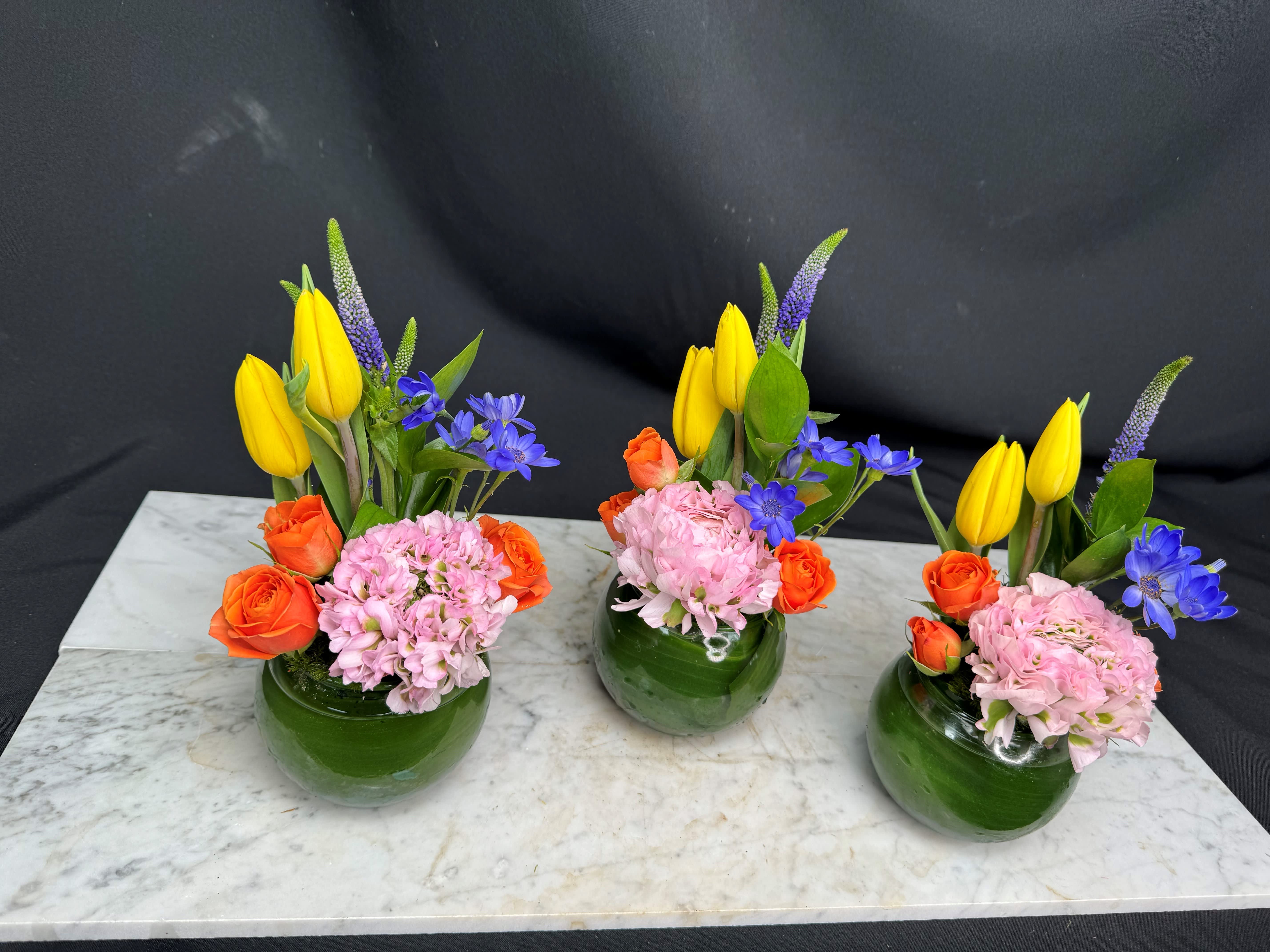 Three small floral arrangements in green glass vases with yellow, orange, pink, and blue flowers
