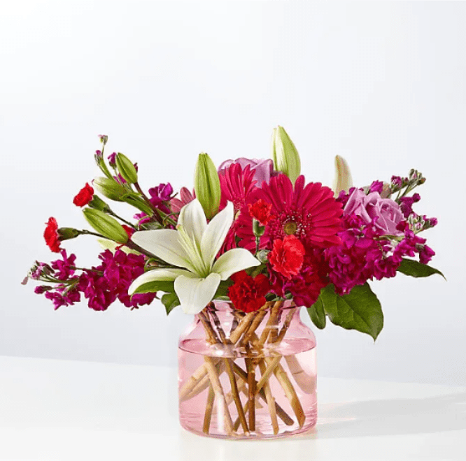 ELEGANT CHARM - Elegant Color, Effortless Charm  This beautifully balanced floral arrangement blends soft romance with vibrant energy, making it a versatile favorite for any occasion. Featuring graceful white lilies, blush and lavender roses, rich red accents, and lively pink blooms, it offers a refined palette with just the right pop of color. Fresh greenery and varied textures add depth, while the modern pink glass vase with exposed stems creates a clean, contemporary presentation.  Soft, elegant color palette with vibrant highlights  Premium blooms chosen for freshness and durability  Professionally designed for balance and fullness  Modern glass vase included, ready to display  Perfect for gifting or enhancing home or office spaces  Chic, timeless, and effortlessly beautiful, this arrangement delivers understated luxury and heartfelt impact—an ideal choice when you want your gift to feel thoughtful, tasteful, and memorable.
