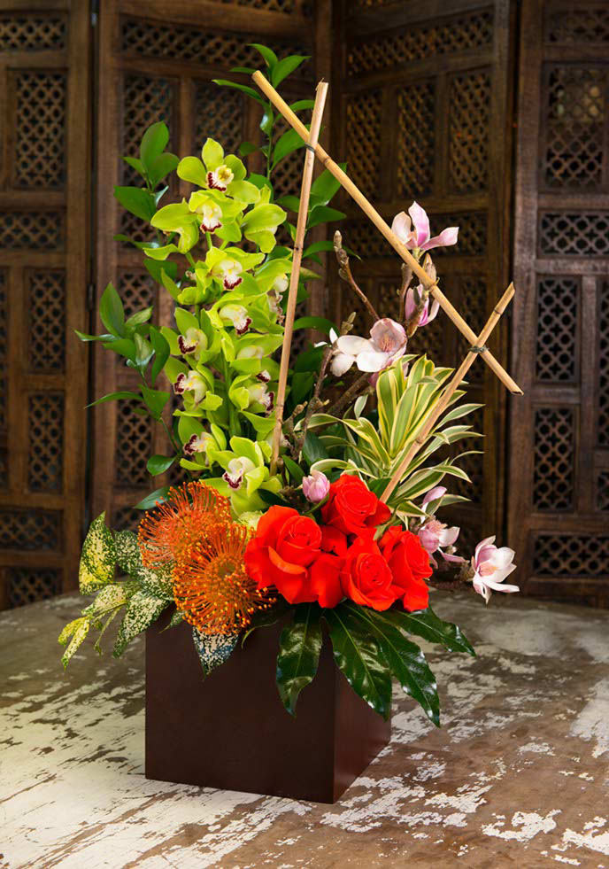 Tall floral arrangement with red roses, green orchids, and orange blooms in a square vase