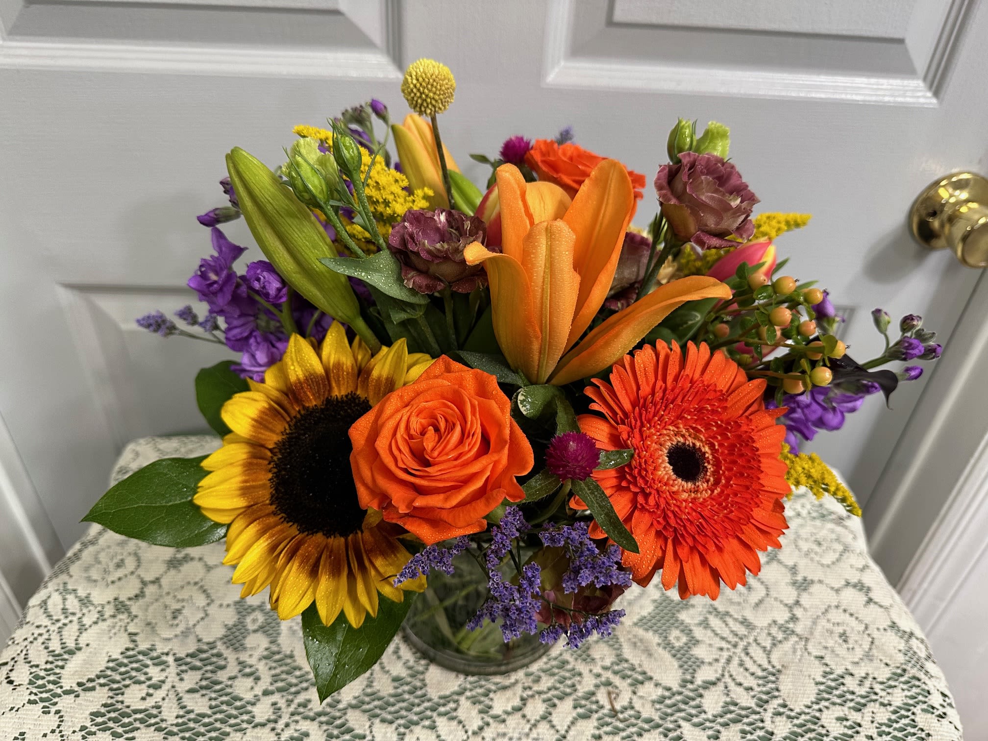 Mixed bouquet with sunflowers, orange roses, lilies, and gerbera daisies in a glass vase