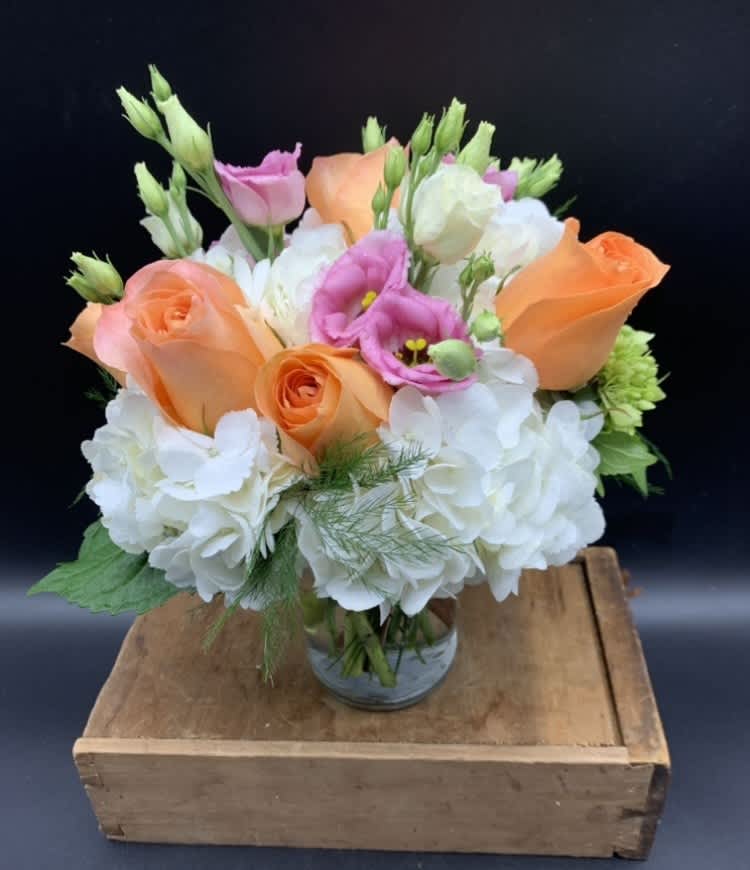 Short arrangement of peach roses, white hydrangeas, and pink lisianthus in a clear glass vase.