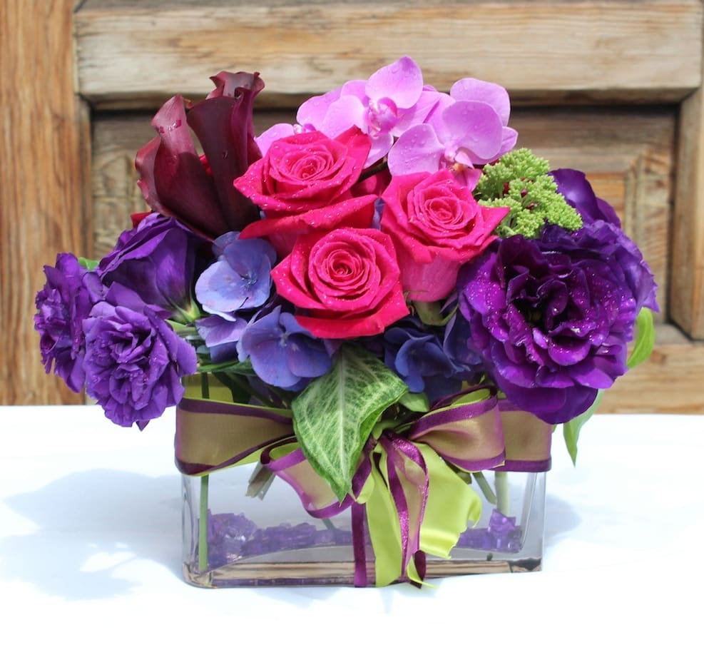Purple and pink floral arrangement in a clear square vase with ribbon