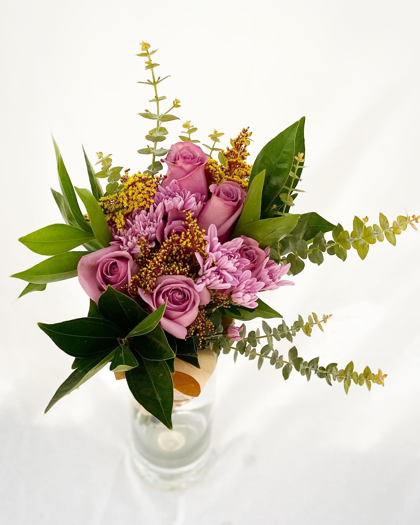 Tall bouquet of mauve roses and lavender mums with yellow filler in a clear glass vase