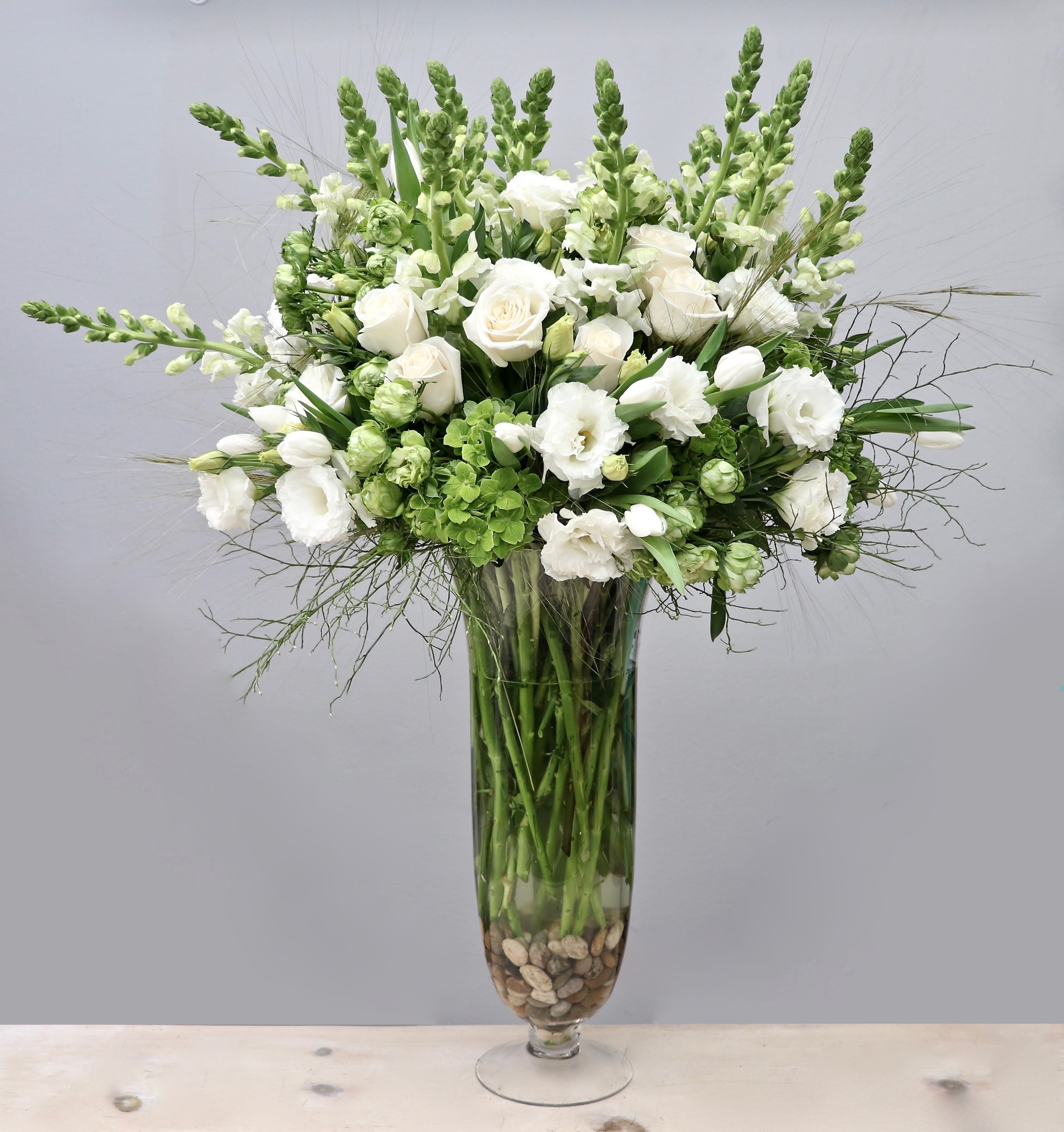 Tall white and green floral arrangement with roses and snapdragons in a clear glass vase with stones