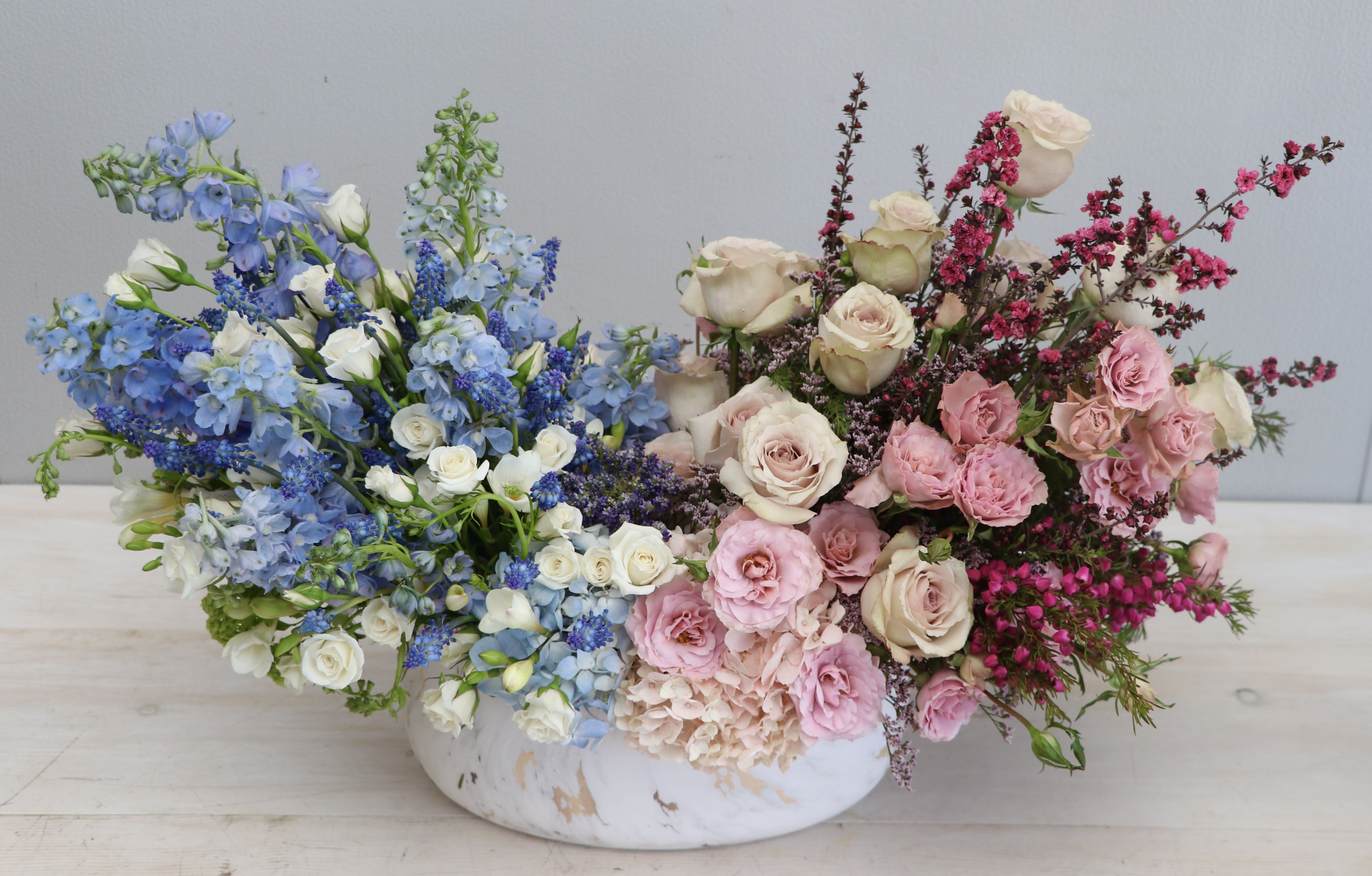 Low centerpiece with blue and white blooms on one side and pink and cream roses on the other in a white bowl