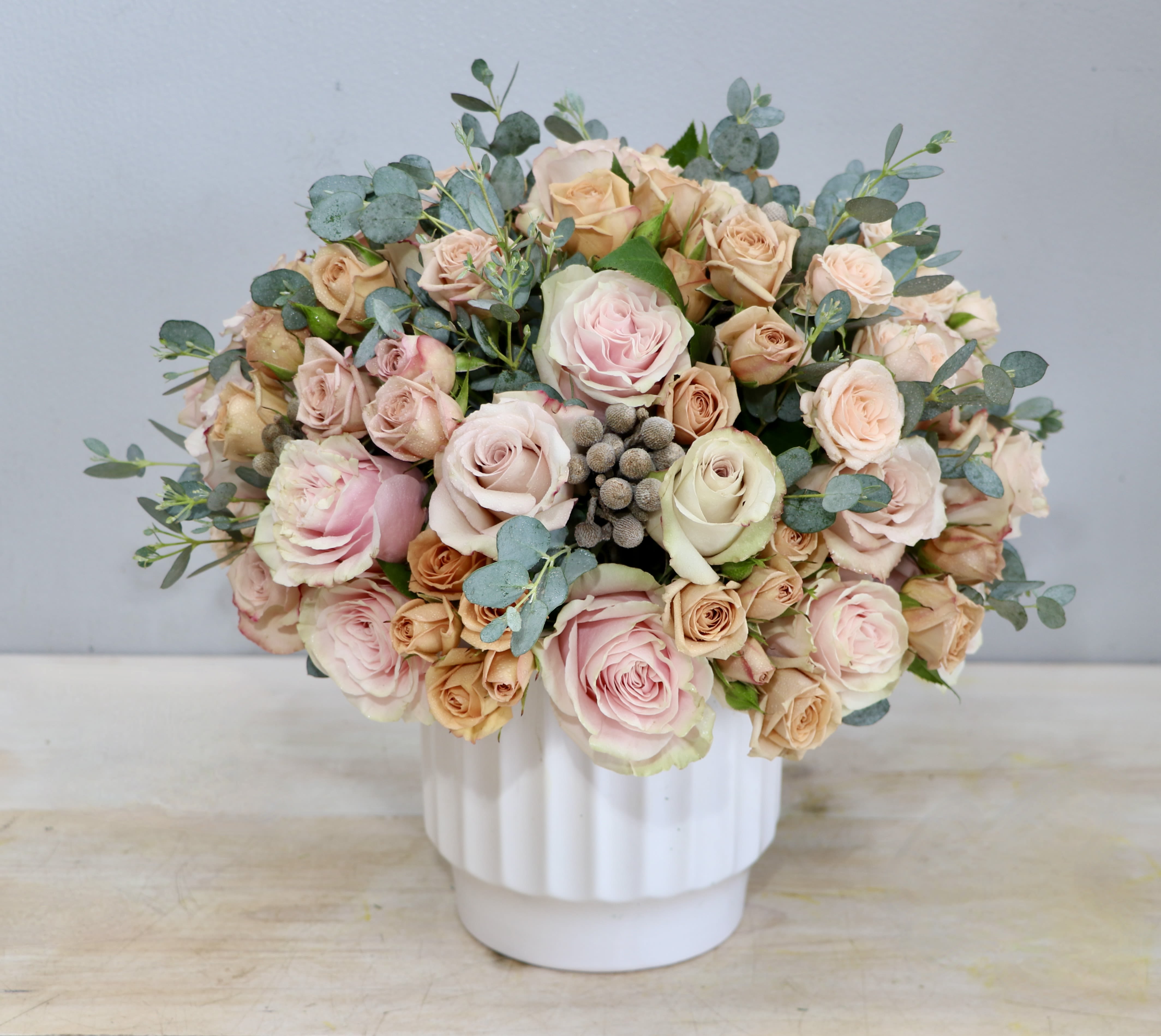 Round arrangement of blush and peach roses in a white ceramic vase with soft gray pods