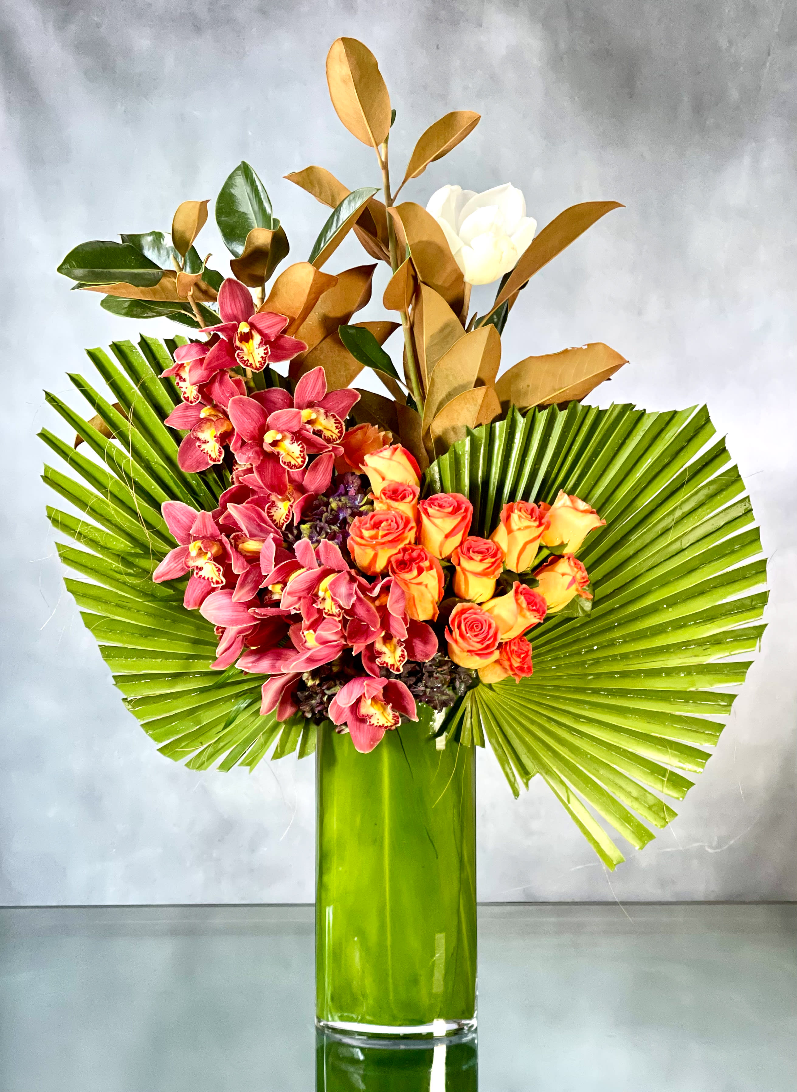 A Dream Come True - Dreams really do come true with this lovely floral composition featuring orange Roses standing tall, surrounded by Cymbidium Orchids and seasonal greens. This stunning composition is presented in a leaf lined tall glass vase filled with fan palms. This arrangement will bring an explosion of color and vibrancy to any room. Approx: 36" H  