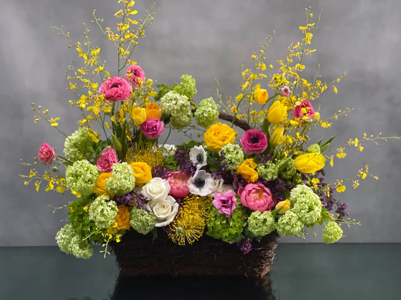 Basket of Spring  - This beautiful basket of happiness will certainly bring a smile to anyone! The basket is filled colorful spring flowers ..... Snowball and green hydrangeas, pink, white and yellow ranunculus, yellow tulips, lilacs, coral peonies, roses, and seasonal florals.  Standard: 22x20 Deluxe : 27x20 ( As Shown)