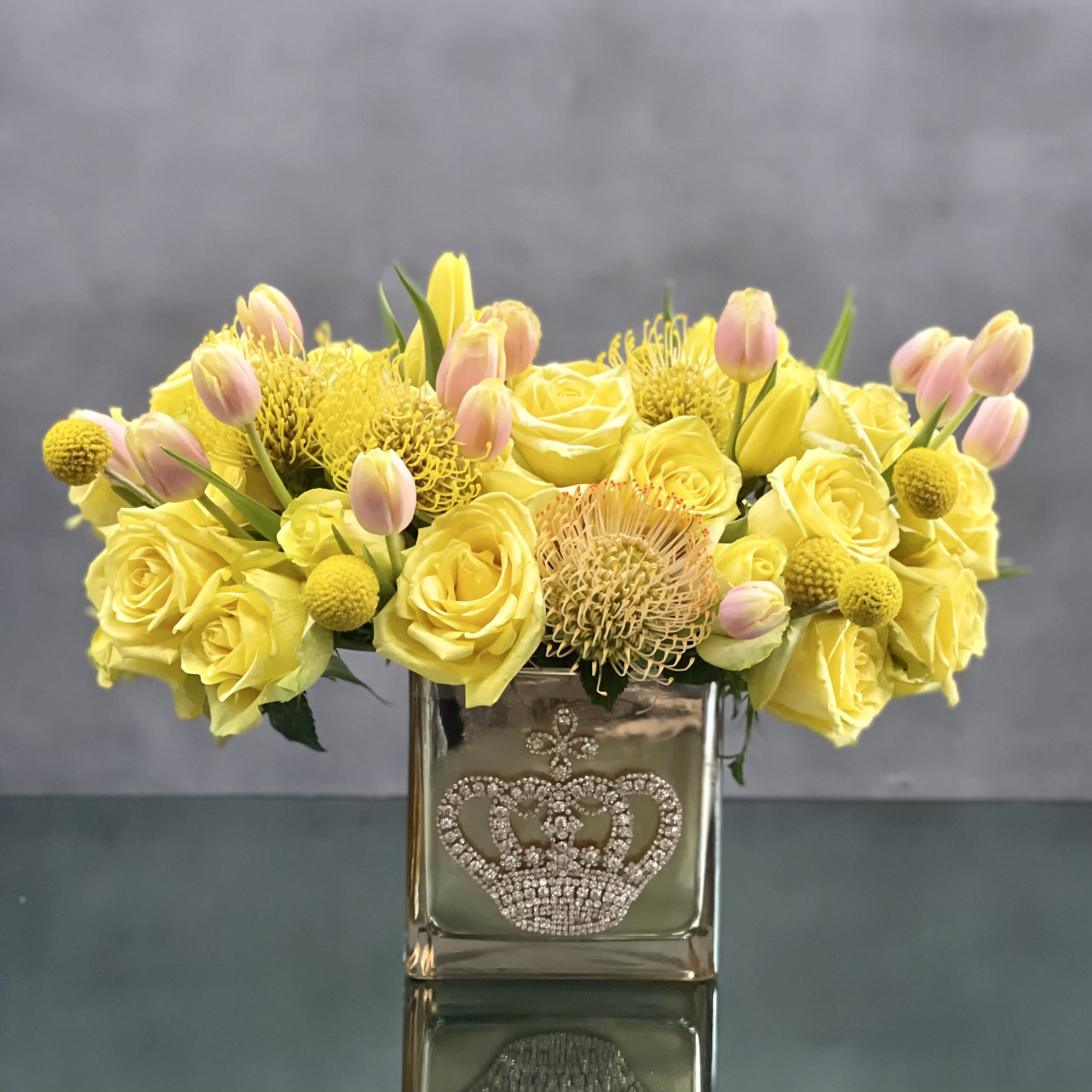 Queen Bee - This striking arrangement of yellow roses, tulips and protea in a gold crown jeweled glass vase. Fit for a queen