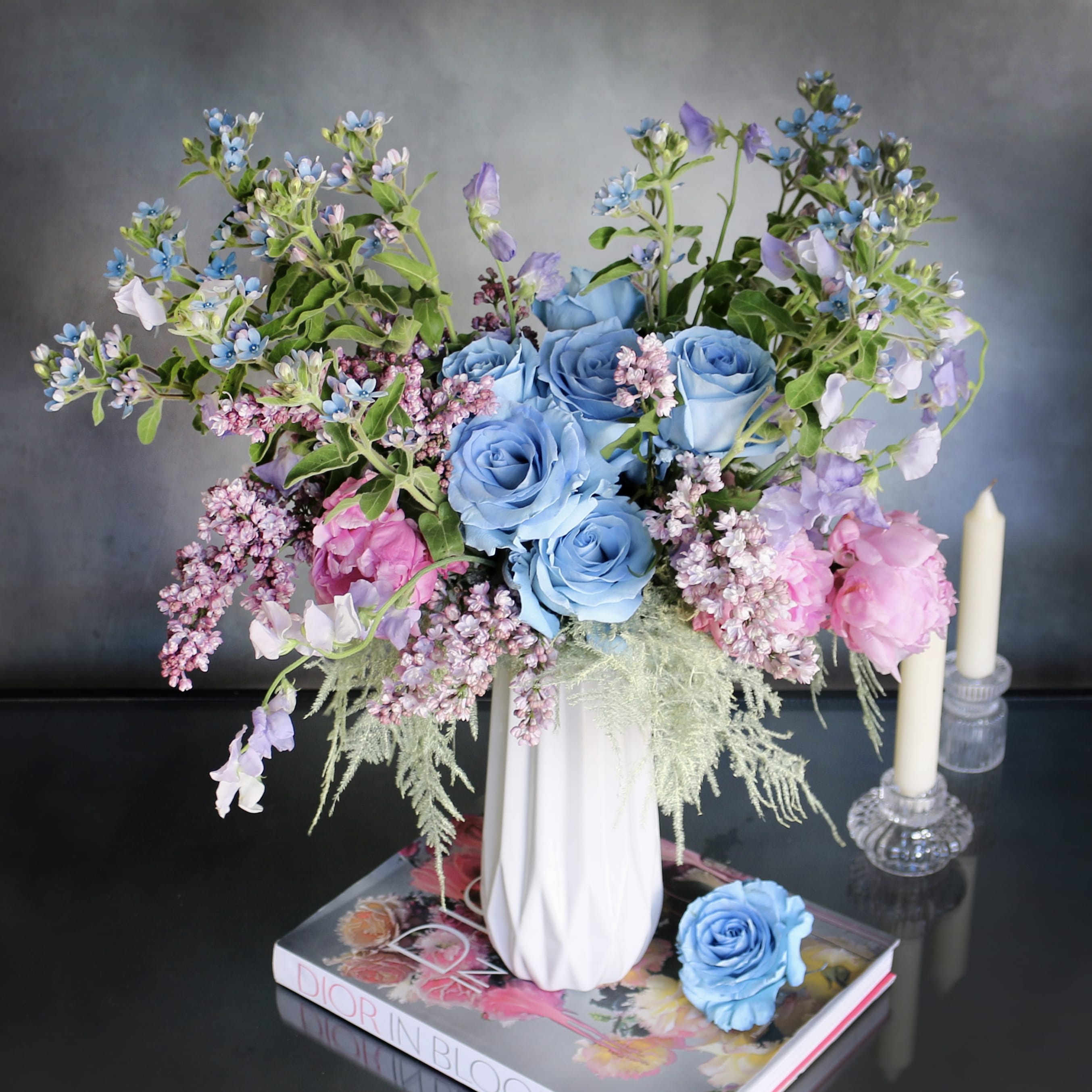 By The Shore  - "By The Shore" consists of blue Roses, Peonies, Sweet Peas, Lilacs and seasonal florals. All arranged within a tall white ceramic vase. A soft and colorful touch to whatever room that the arrangement is placed in. A bright focus for any occasion.   Measurement:  Vase: 11 x 5 Arrangement: 23 x 15