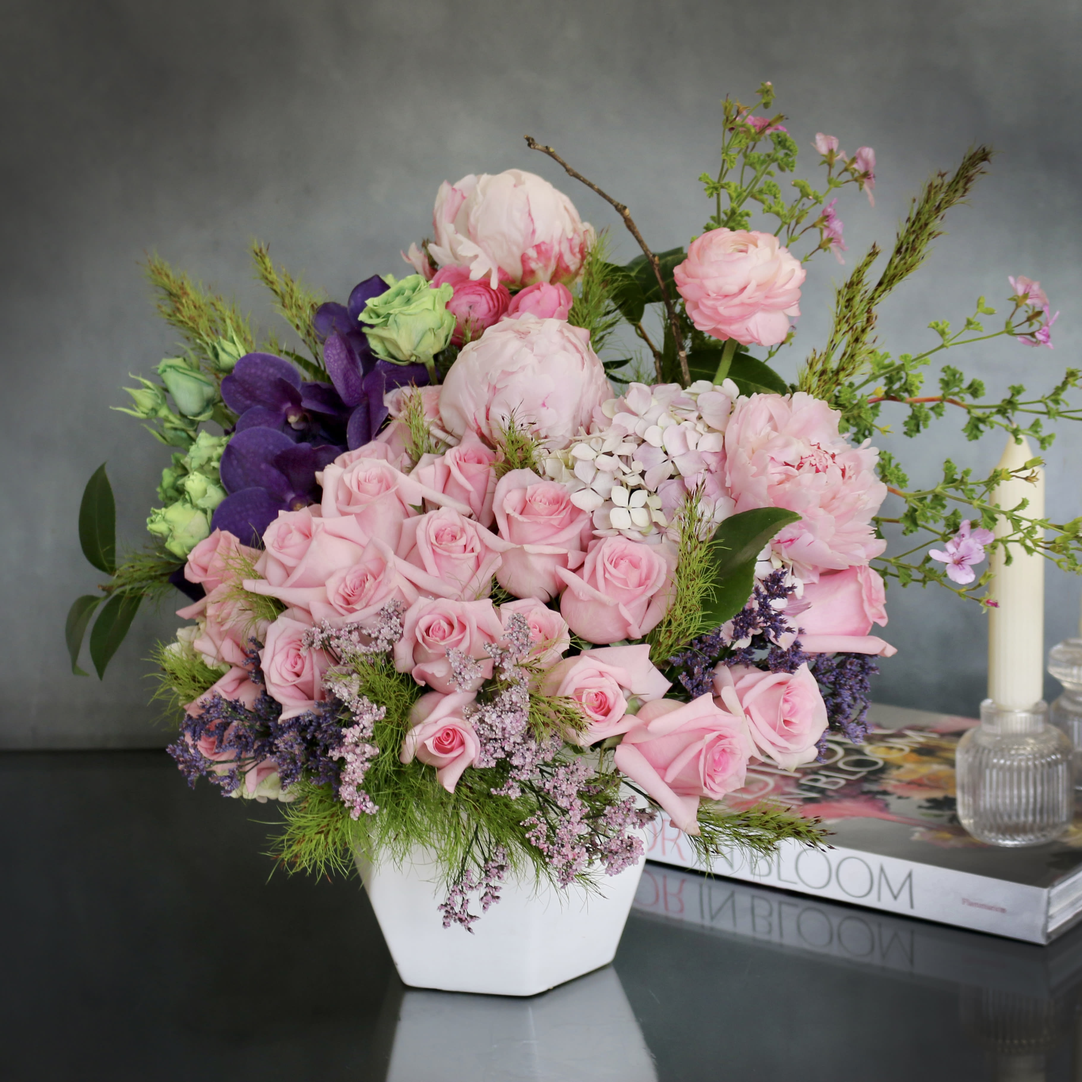 Floral Escape  - Radiating beauty and elegance, this delicate and dainty floral design features a soft palette of pink with a touch of purple, creating a charming visual delight. Adorned with lovely Vanda orchids, stunning roses, and peonies alongside other seasonal flowers, this arrangement paints a beautiful floral picture that exudes grace and sophistication, all nestled inside a white ceramic vase.  In the premium size variation, this arrangement boasts 25 roses and peonies, ensuring a lavish and opulent display. Opting for the standard size will include fewer flowers, with a focus on either peonies or orchids to maintain the essence and charm of the arrangement while adjusting for a variation in floral components.