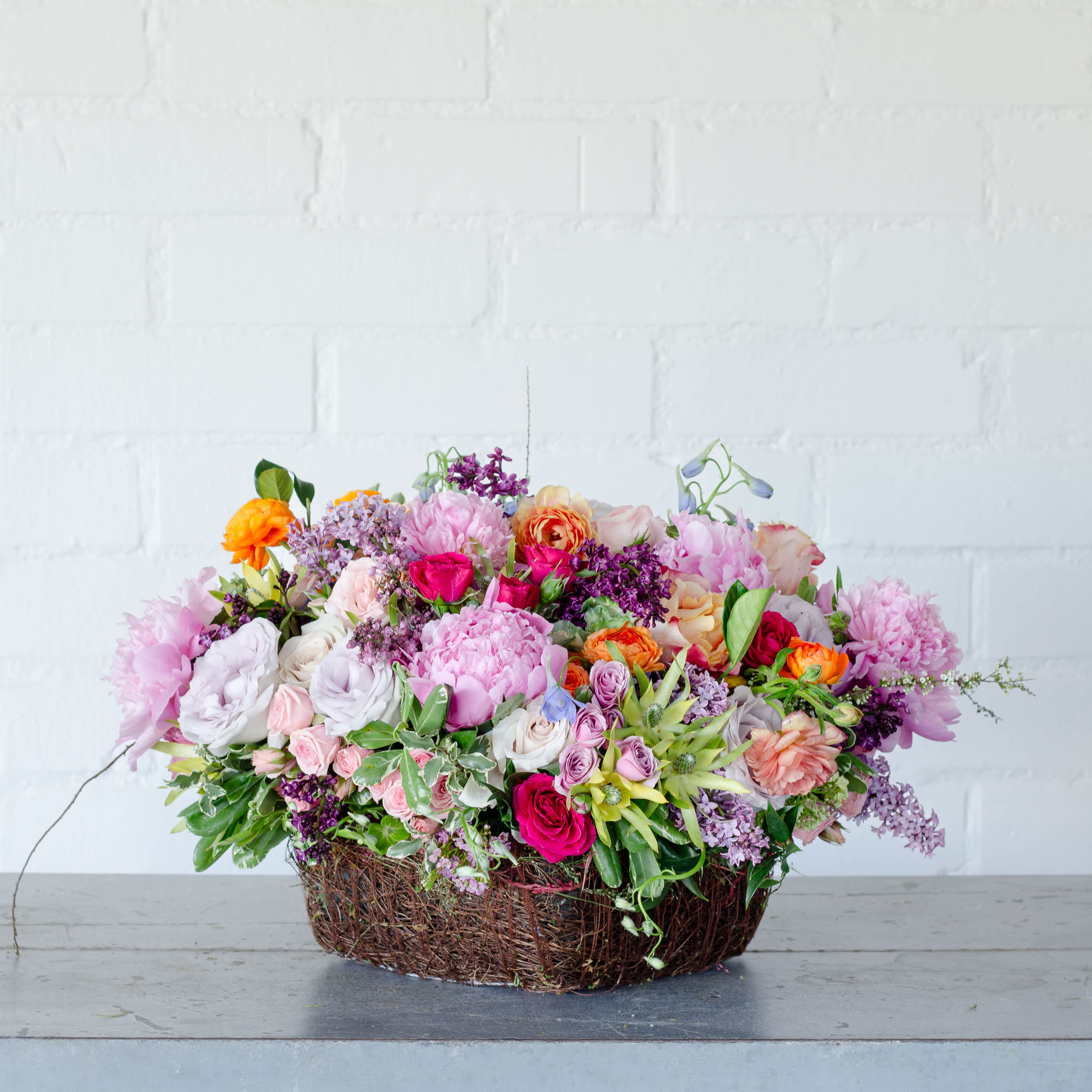 GARDEN MEDLEY - Same day delivery from your local florist!   a stunning floral arrangement fitting for Mother's Day, available for same-day delivery through a local florist, could include garden roses, peonies, lilacs, and other local flowers beautifully arranged in a tall square basket filled with fresh oranges. This combination of blooms offers a delightful array of colors, scents, and textures, creating a perfect gift to honor and appreciate a special mother figure.  The floral arrangement would be approximately 16 inches wide and 7 inches tall, showcasing the vibrant beauty of the garden roses, the lushness of peonies, the fragrant lilacs, and the unique charm of local flowers. This arrangement is sure to bring joy and warmth to any Mother's Day celebration.