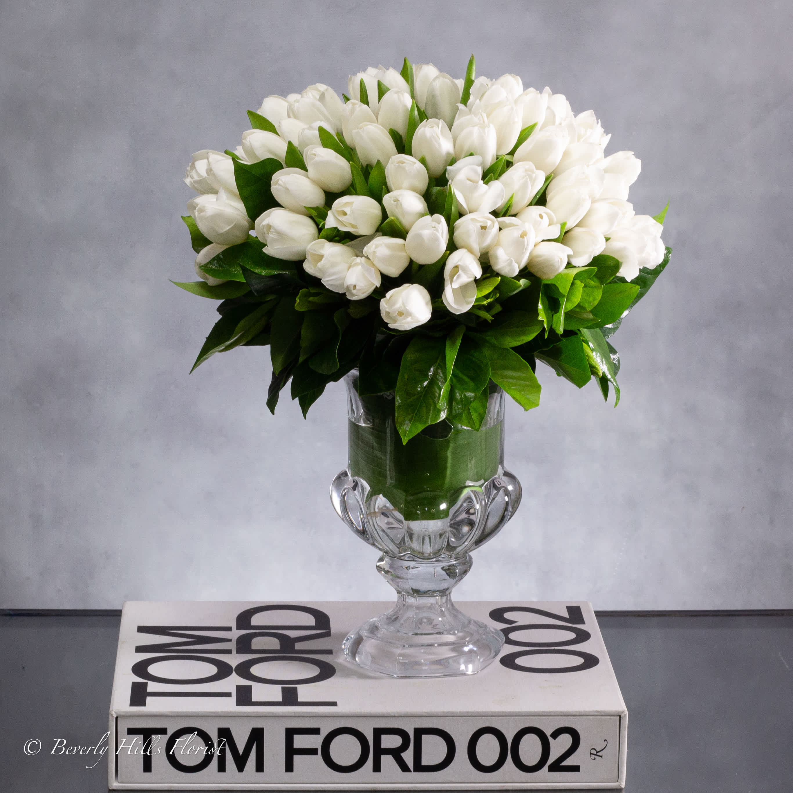 WHITE TULIPS IN CRYSTAL SPLENDOR - White tulips symbolize purity, innocence, and new beginnings. When arranged in a white crystal urn, these symbols are enhanced by the elegance and sophistication of the setting.  The 8" crystal urn adds a layer of purity and refinement to the 14" tall arrangement, reflecting the pristine nature of the tulips themselves. The crystal's transparency allows the beauty of the tulips to shine through, emphasizing their purity and simplicity.  Together, the white tulips and the white crystal urn create a harmonious composition that conveys a sense of serenity and tranquility. It's a timeless and elegant arrangement that speaks of new beginnings, making it perfect for weddings, celebrations, or simply as a gesture of hope and renewal.