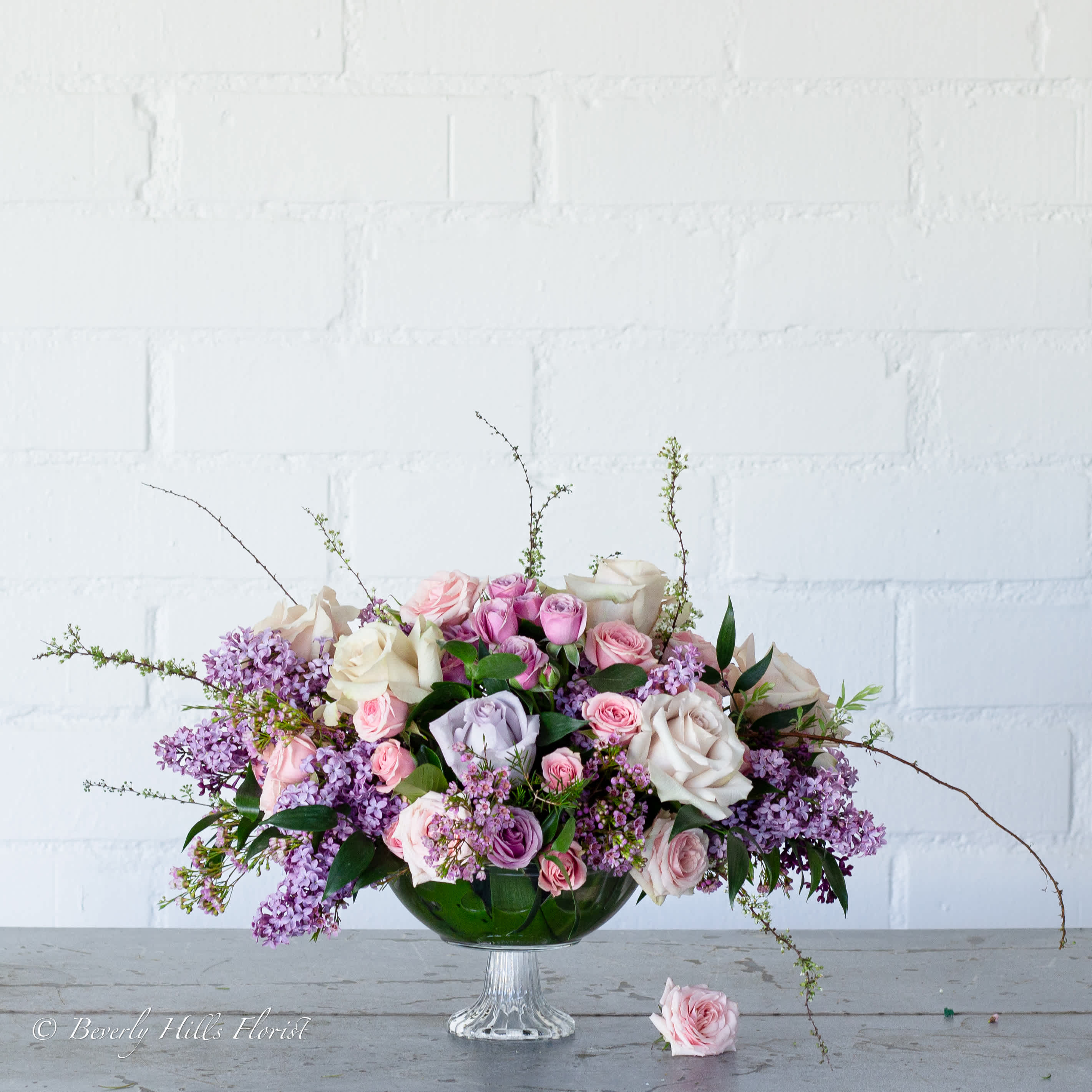 A Symphony of Lilacs and Roses - A beautiful floral arrangement featuring local lilacs and soft-colored roses set in a pedestal glass vase. The lilacs add a delicate touch with their light hues and unique fragrance, while the soft-colored roses complement them perfectly. The arrangement is 18 inches wide and 12 inches tall, creating a lovely and elegant centerpiece for any space. Placing it in a pedestal glass vase adds a touch of sophistication and allows the flowers to shine.