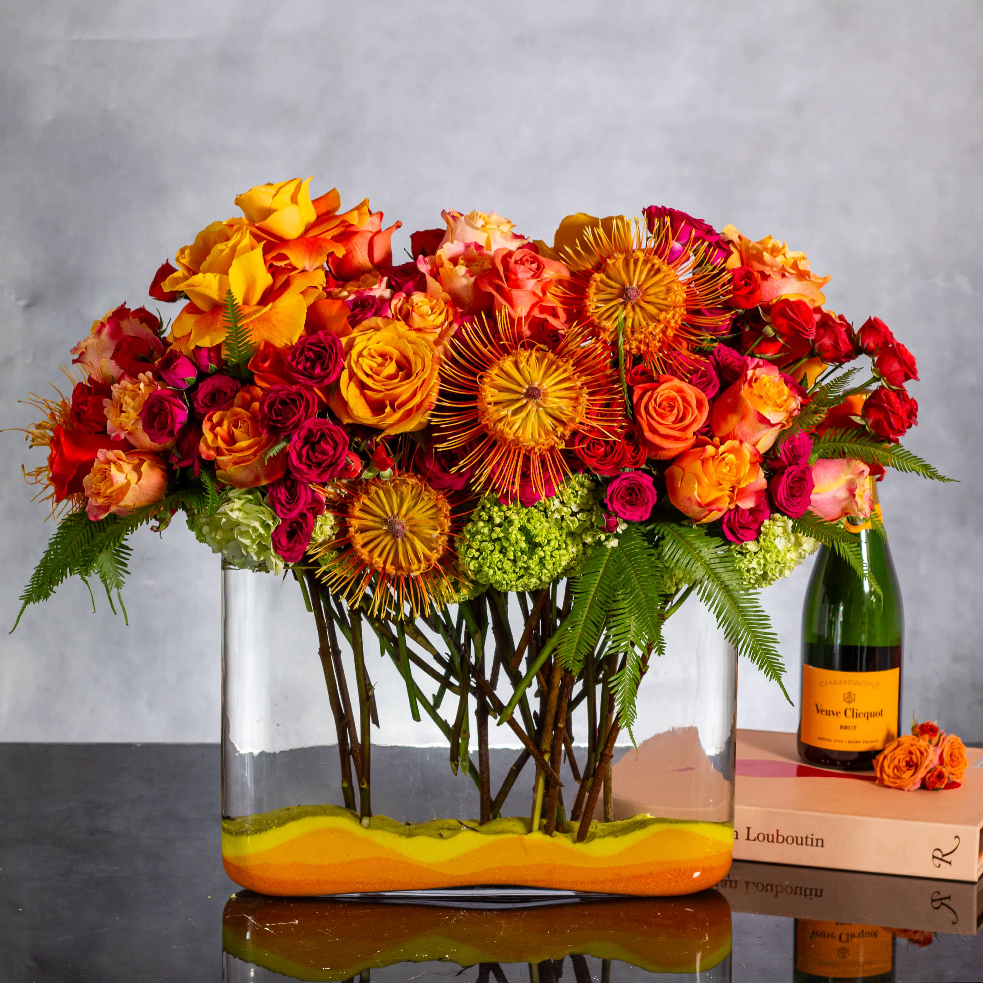 Sunset beauty - Indulge in a vibrant display reminiscent of a majestic tropical sunset with this enchanting arrangement featuring orange and yellow roses, delicate orange orchids, and exotic tropical blooms. The harmonious blend of colors creates a stunning visual akin to a mesmerizing sunset sky.  Complete in an 18-inch long and about 22-inch tall arrangement, this floral ensemble exudes warmth and sophistication. The combination of fiery oranges and sunny yellows, accentuated by the tropical beauty of the orchids and other blooms, transports you to a paradise of vibrant hues and natural splendor.