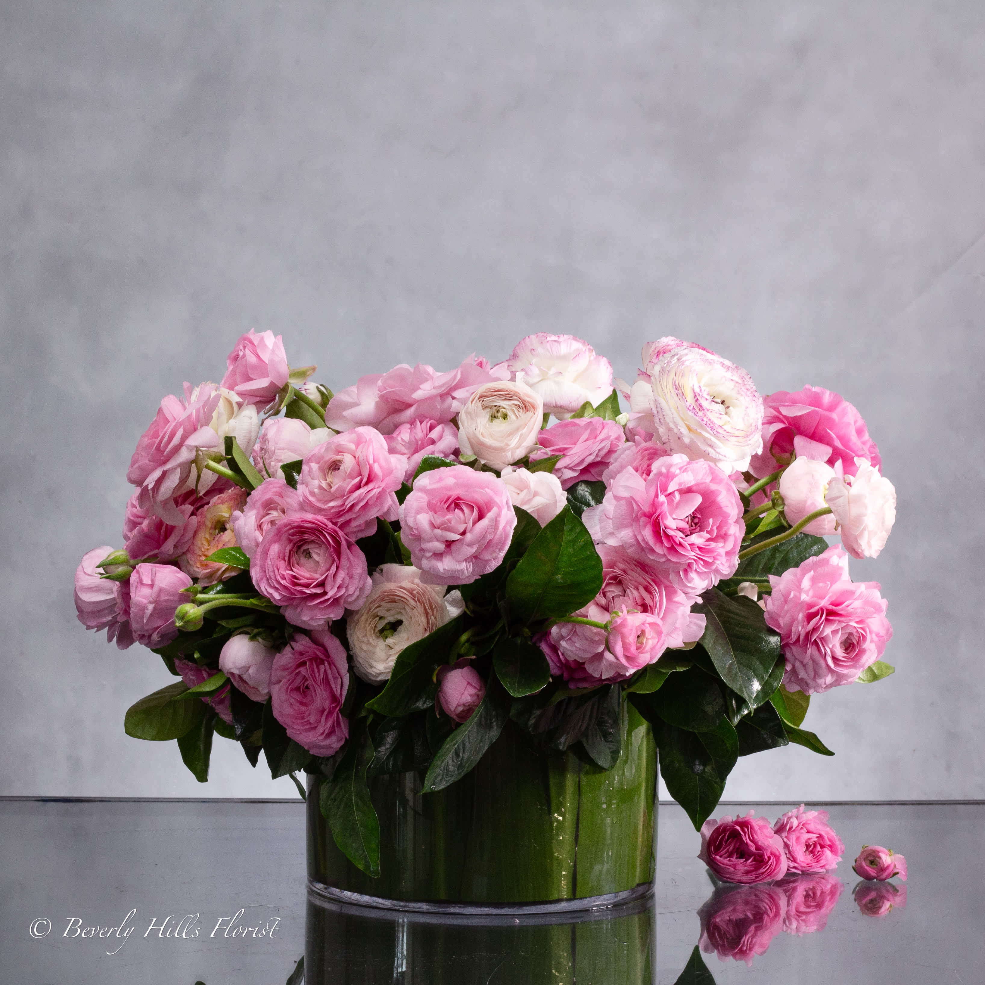 MODERN ROMANCE: PINK RANUNCULUS IN A CYLINDER OF GRACE - Pasadena Florist - Pink ranunculus symbolize charm, attractiveness, and admiration. When arranged in a cylindrical glass vase, these meanings are enhanced by the contemporary and transparent nature of the vessel.  The pink hue of the ranunculus signifies admiration and appreciation, making them an ideal choice for expressing fondness and admiration towards someone. The cylindrical glass vase provides a modern and sleek backdrop for the delicate beauty of the ranunculus, allowing their petals to be showcased in all their glory.  The transparency of the 8" glass vase adds a sense of openness and clarity to the 13" overall round arrangement, symbolizing sincerity and transparency in emotions. It also allows for the stems of the ranunculus to be visible, adding to the overall aesthetic appeal of the display.  Together, the pink ranunculus and cylindrical glass vase create a sophisticated and contemporary floral arrangement that is both visually striking and meaningful. It's a perfect choice for adding a touch of charm and elegance to any space or occasion.