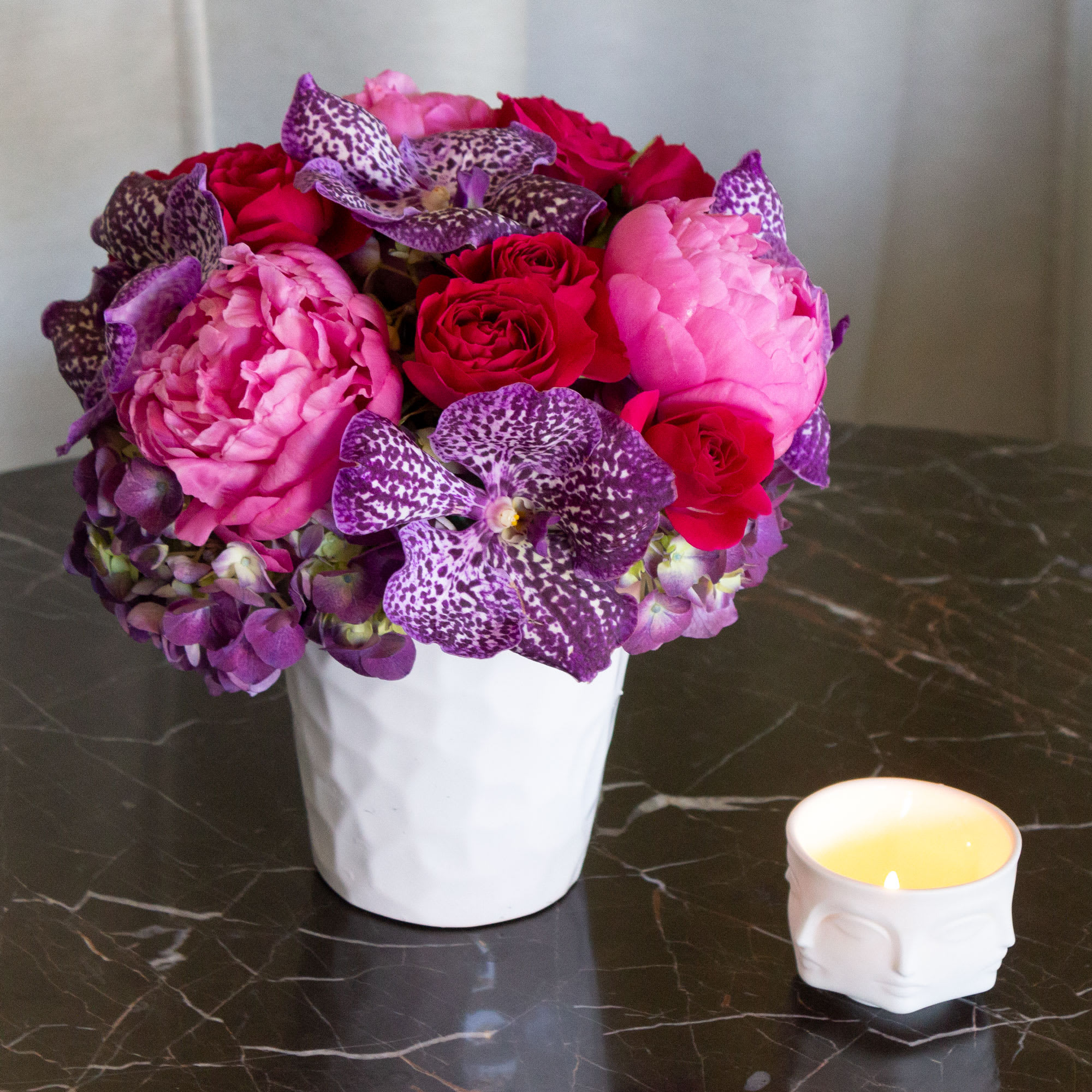 Portrait of Peonies - Indulge in a bouquet that is both good and girly, designed to captivate with its trendy flair. This exquisite arrangement combines the timeless allure of Roses and pink Peonies with the exotic charm of purple Vanda Orchids and the lushness of hydrangeas. Each bloom is carefully selected to create a harmonious blend of colors and textures, perfect for the modern feminine aesthetic.  Presented in a decorative white ceramic pot, this arrangement exudes sophistication and style, adding a touch of elegance to any space. Whether it's a birthday, anniversary, or just a special surprise, this floral masterpiece is sure to delight and enchant. With an approximate size of 10" round, the Enchanted Bloom is a beautiful statement piece that will leave a lasting impression."