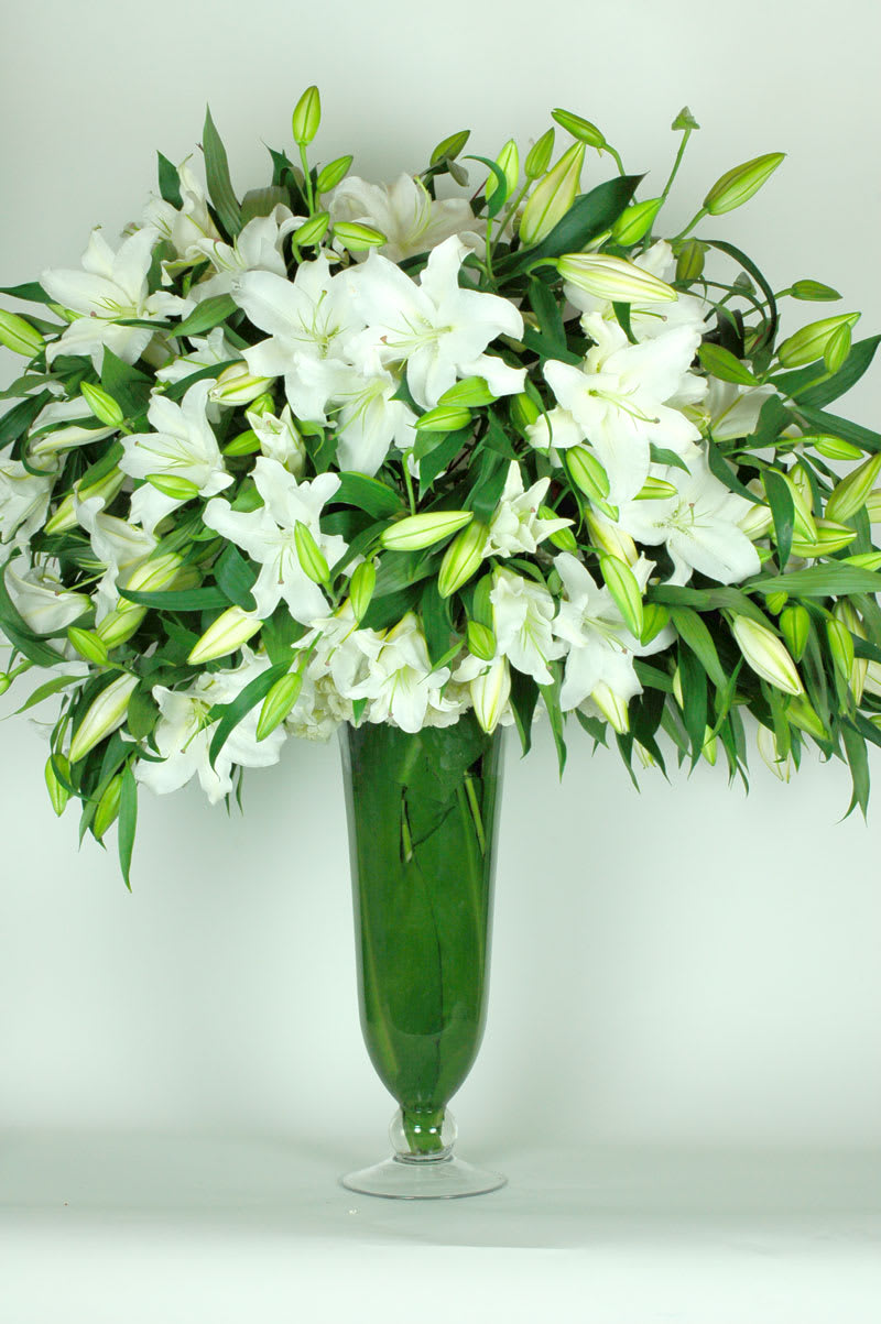 Tall arrangement of white lilies and buds in a slender green glass vase