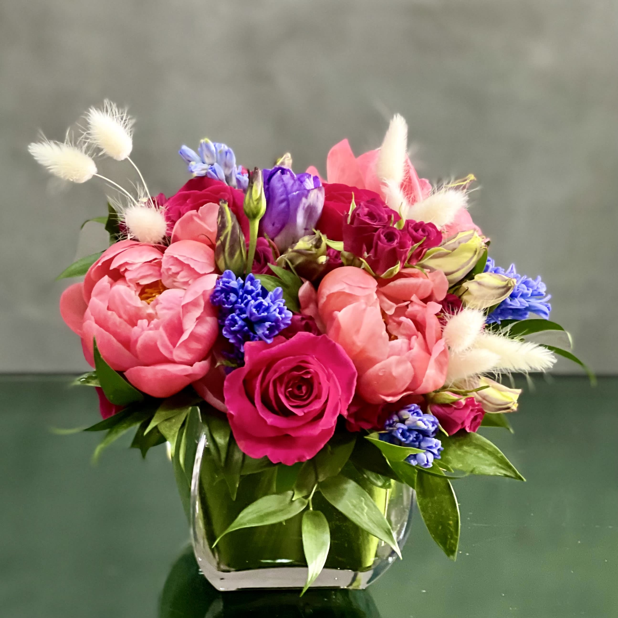 Posh Peonies - This striking combination of large full coral Peonies, contrasted with the deep tones of Lisianthus and clusters of spray Roses creates a unique eye-catching design that stands above the rest.    Base is 4x4 glass cube . Overrall arrangement is around 8" inches   Standard as shown