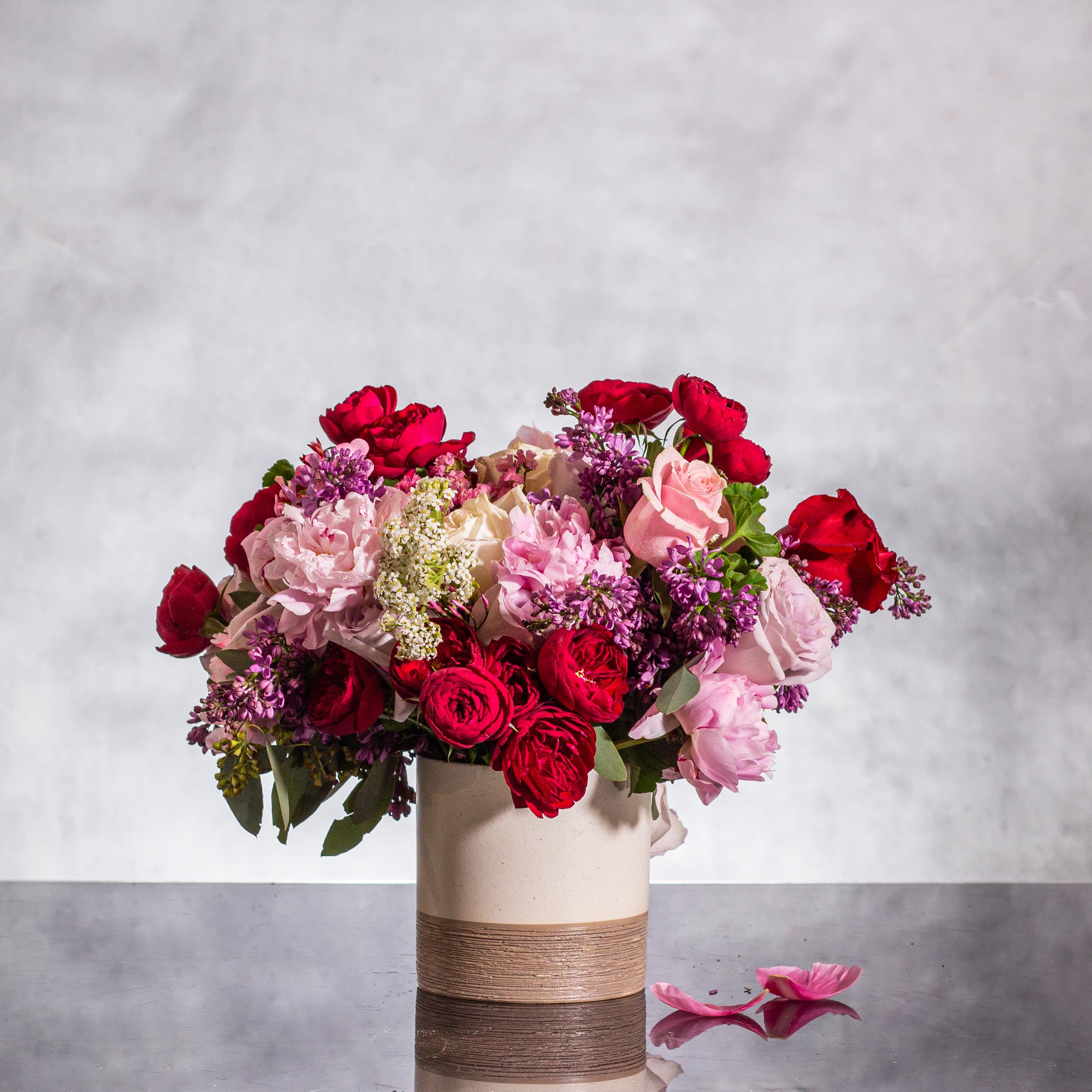 Introducing our Seductive Blooms - Immerse yourself in a world of seduction with our enchanting floral creation. Picture clusters of deep burgundy garden roses entwined with the intoxicating scent of luscious lilacs, all housed in a beautifully arresting ceramic base standing at approximately 15 inches tall and 12 inches wide. This arrangement exudes an irresistible allure, weaving a tale of passion and desire with every petal. Indulge in the sensual beauty of these blooms, creating an ambiance of pure seduction in any setting.