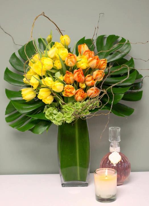 Tulip Tango - Spectacular and dramatic ! This knock-out design is nothing short of a Tropical Paradise!  Gorgeous clusters of radiant sunny yellow and orange Tulips burst from the large leafy greenery background standing tall in a elegant glass leaf-lined vase. A refreshing surprise that celebrates the bright side of life!