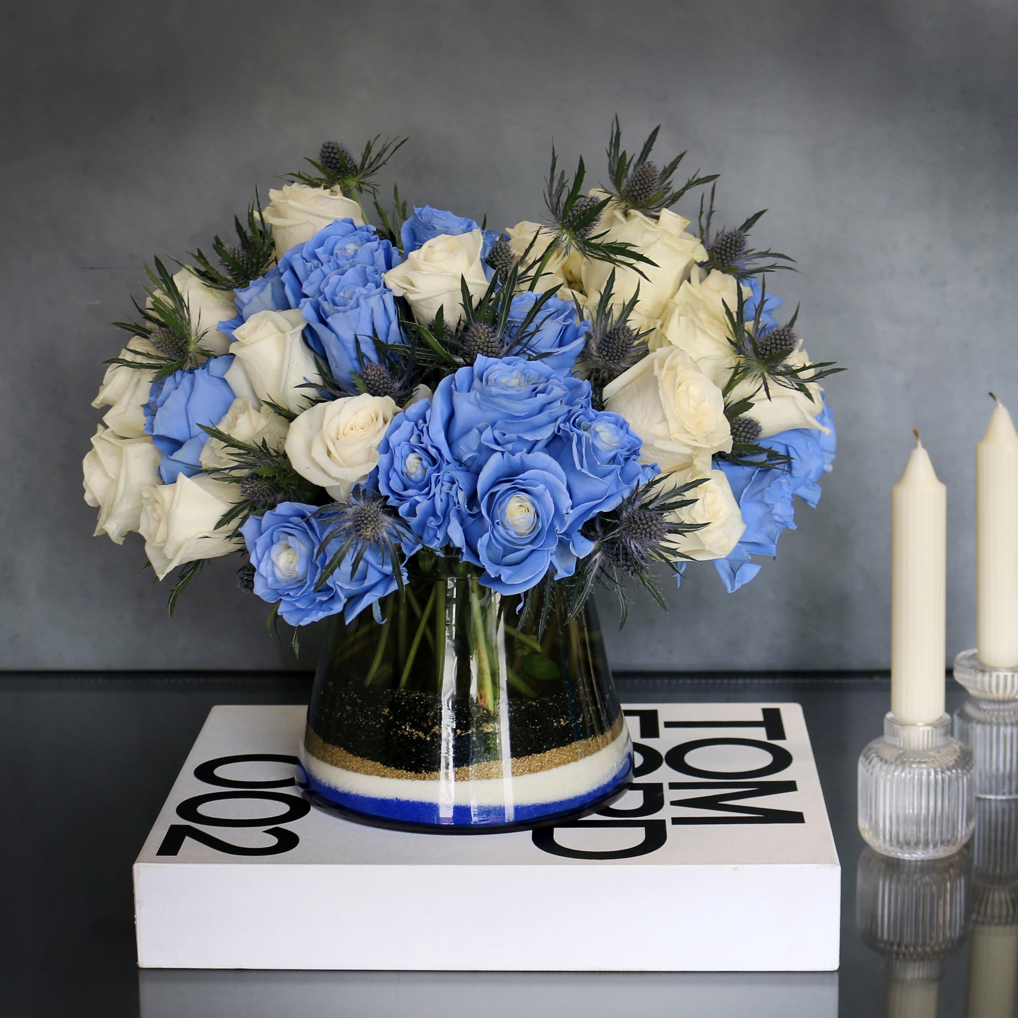 Blue Me Away  - "Blue Me Away" is a unique new twist to a simple vase arrangement. The arrangement consists of blue and white Roses and Thistles. Which is then accented with various colors of sand at the bottom of the vase.   Measurement:  Vase: 7x8 Arrangement: 14x16