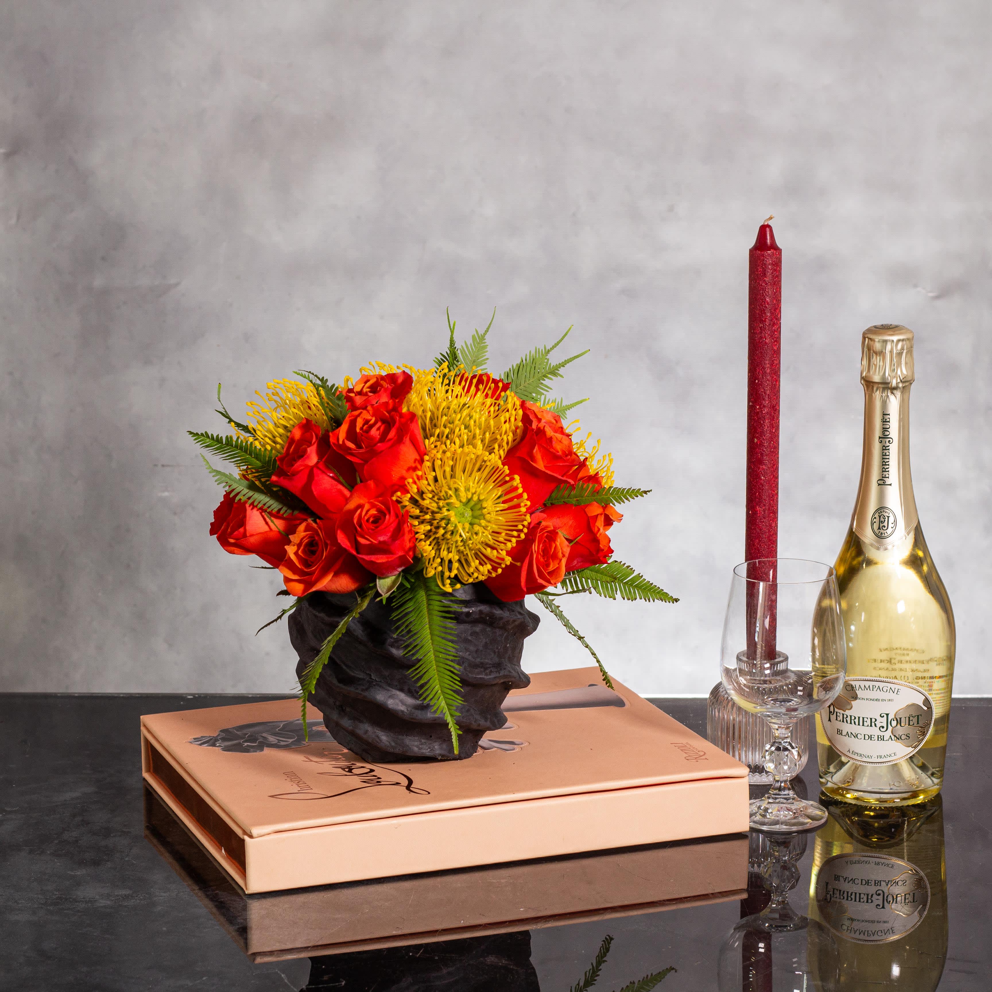 Vibrant Citrus Delight - Experience a burst of color with this vibrant arrangement featuring bright orange roses and sunny yellow proteas, elegantly displayed in a sleek black modern container vase. The striking contrast between the orange roses and yellow proteas creates a lively and cheerful composition that is guaranteed to captivate.  Nestled in a chic black modern container vase, approximately 9 inches round, this arrangement exudes a contemporary flair that perfectly complements the bold colors of the flowers. The modern and sophisticated design of this floral ensemble makes it an eye-catching centerpiece for any space, adding a touch of elegance and vibrancy.  Whether gracing a dining table, enhancing a living room, or delighting as a thoughtful gift, this arrangement of orange roses and yellow proteas in a black modern container vase is a perfect choice for those seeking a stylish and colorful floral display.