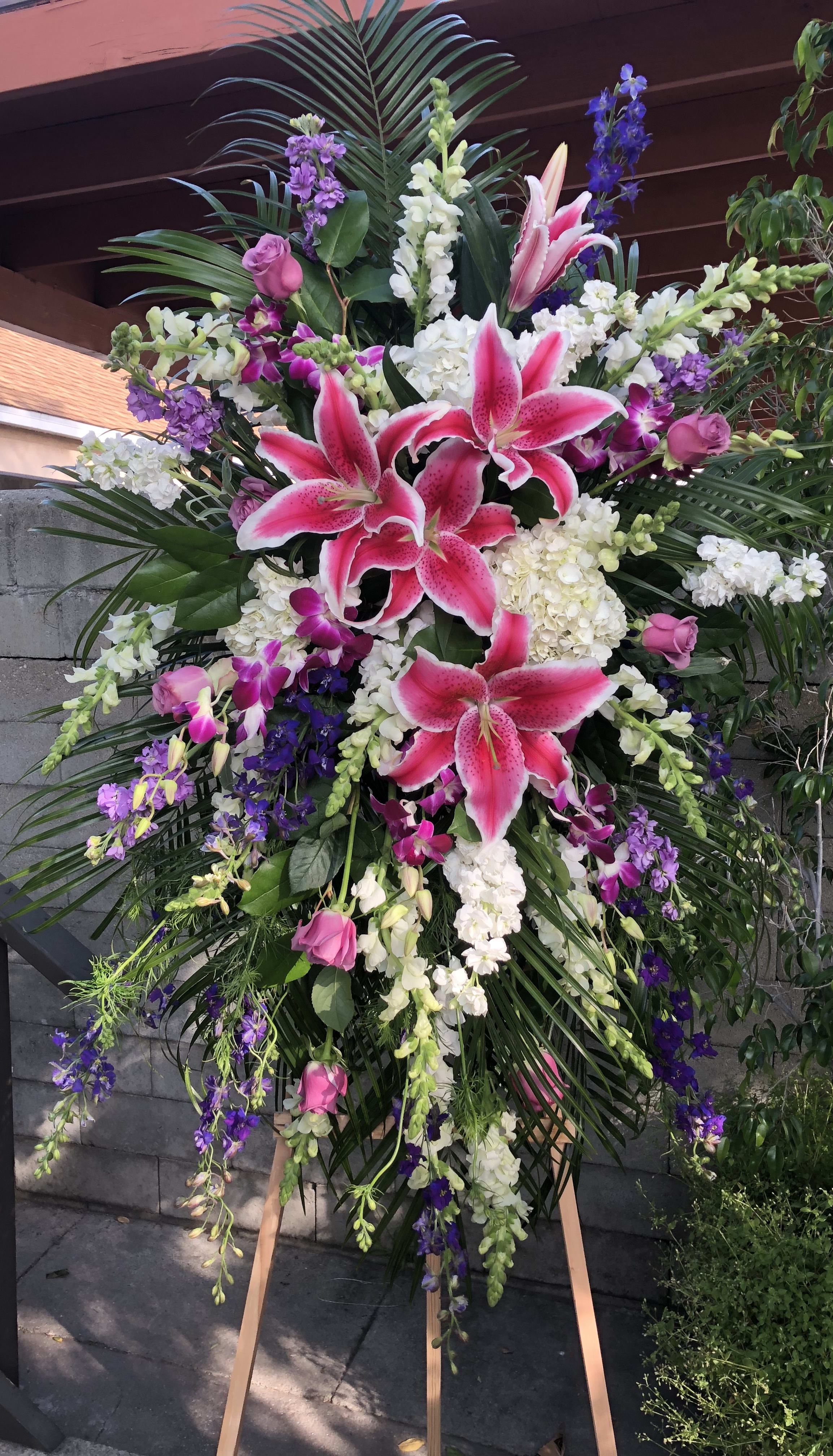 Tall standing spray with pink lilies, white flowers, purple blooms, and palm fronds on a wooden easel
