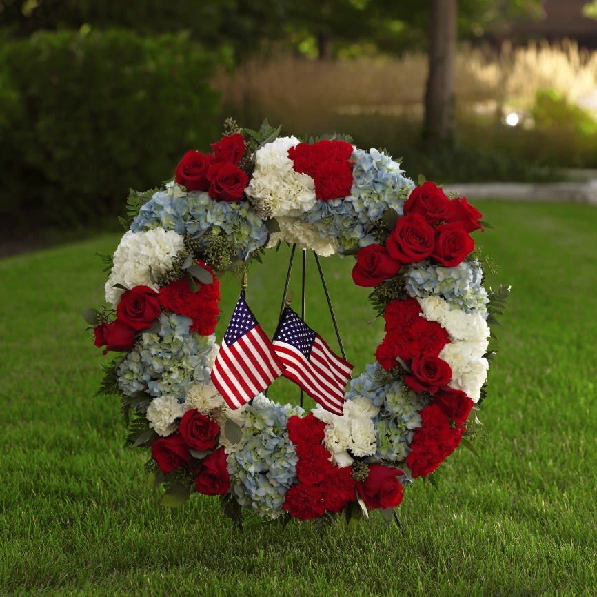 Floral wreath with red roses, white carnations, blue hydrangeas, and two small American flags