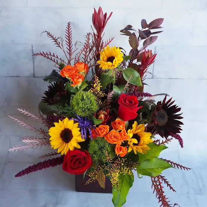 Mixed bouquet with sunflowers, roses, and orange spray roses in a square vase