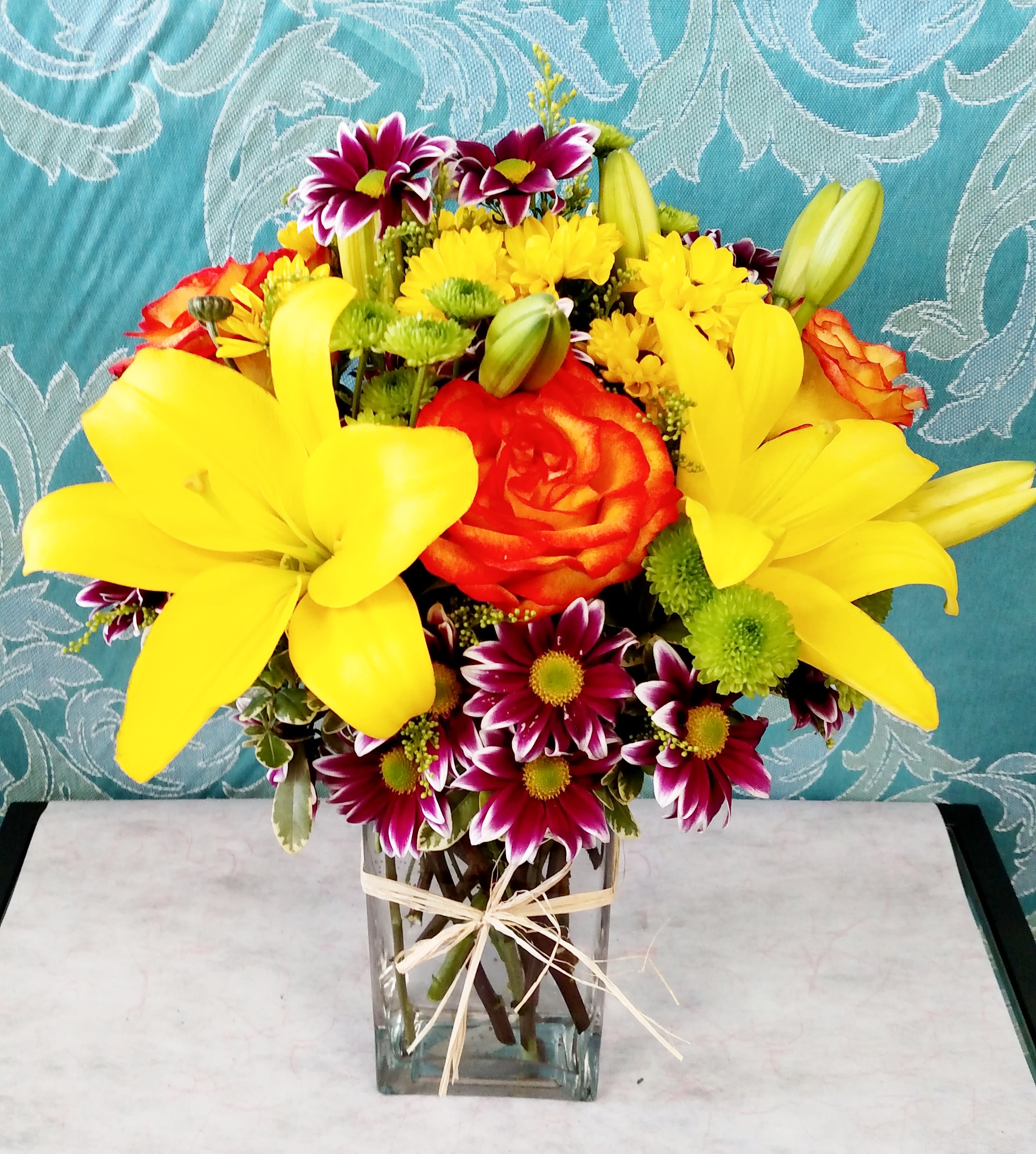 Bright bouquet of yellow lilies, orange roses, and mixed daisies in a clear rectangular vase