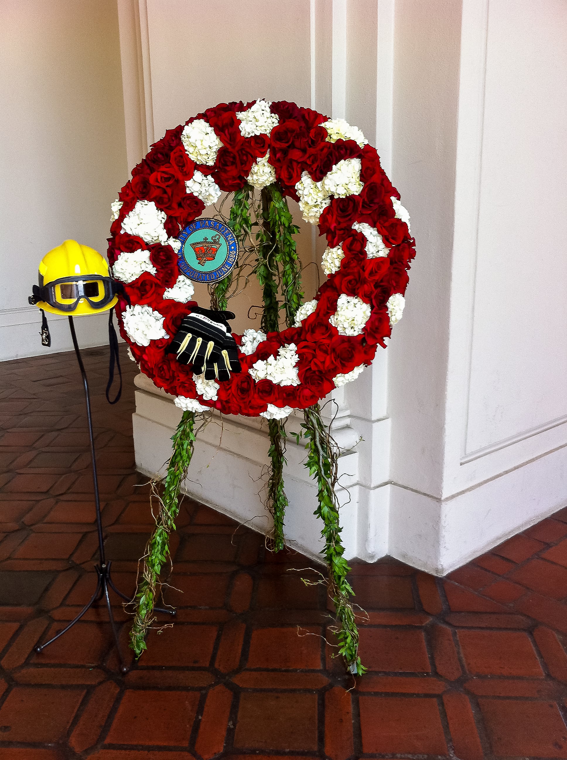 Red and white floral wreath on an easel with a firefighter helmet and gloves