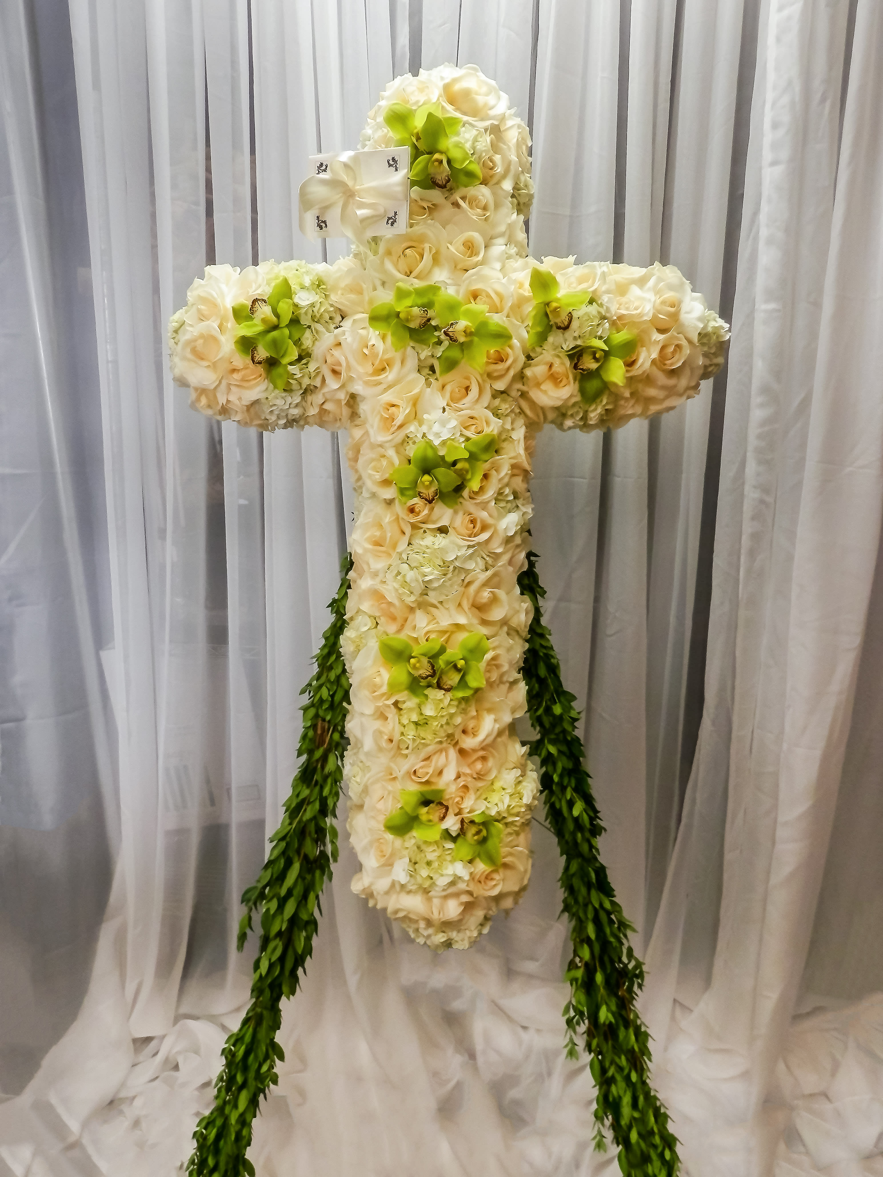 Cross-shaped floral arrangement of cream roses and green orchids with trailing greenery