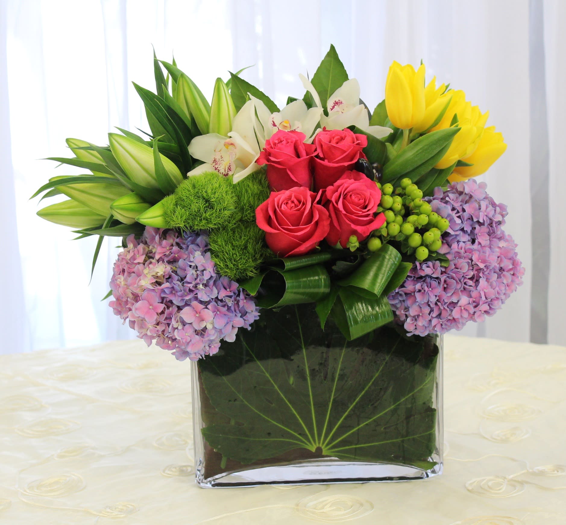 Mixed bouquet in a clear square vase with roses, lilies, hydrangeas, and tulips