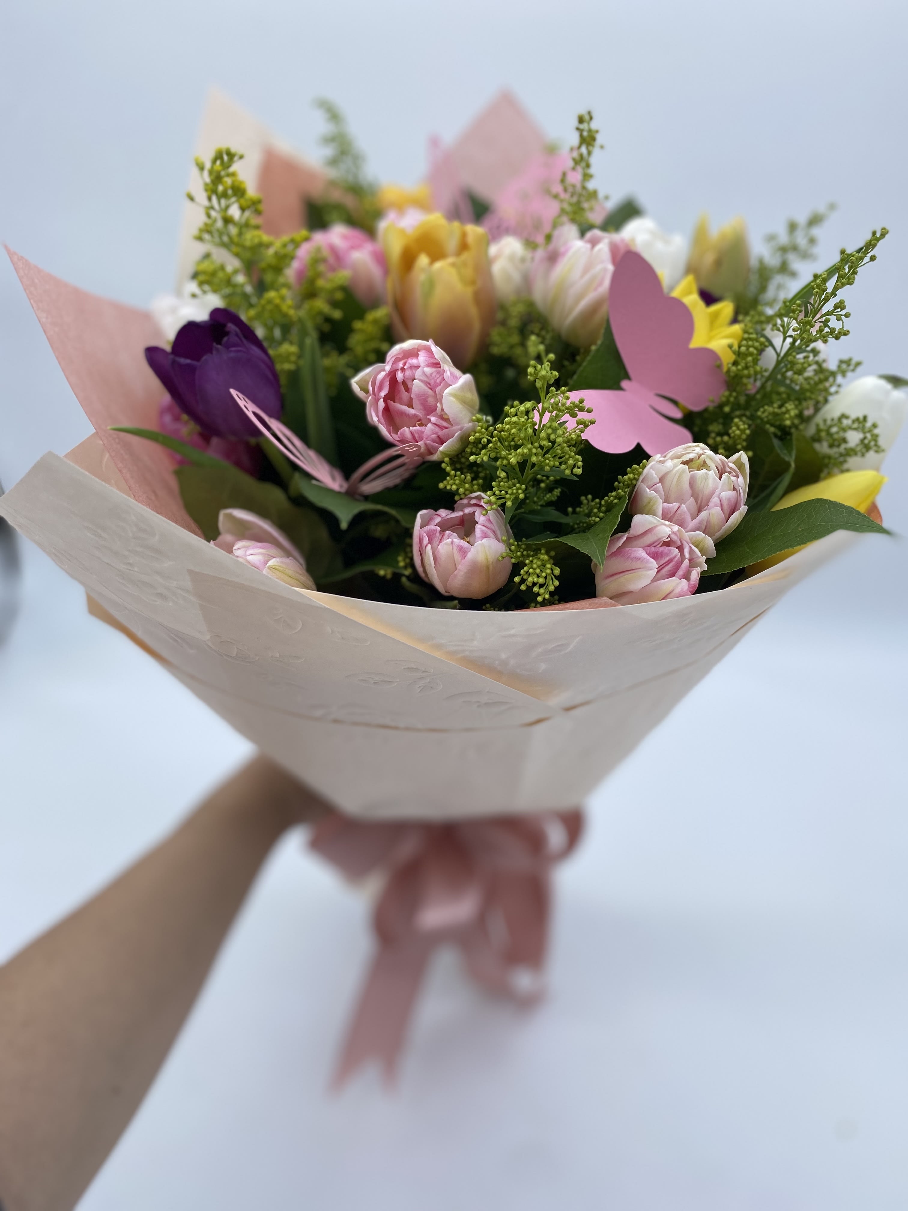 Handheld bouquet of pink, yellow, and purple tulips wrapped in pale paper