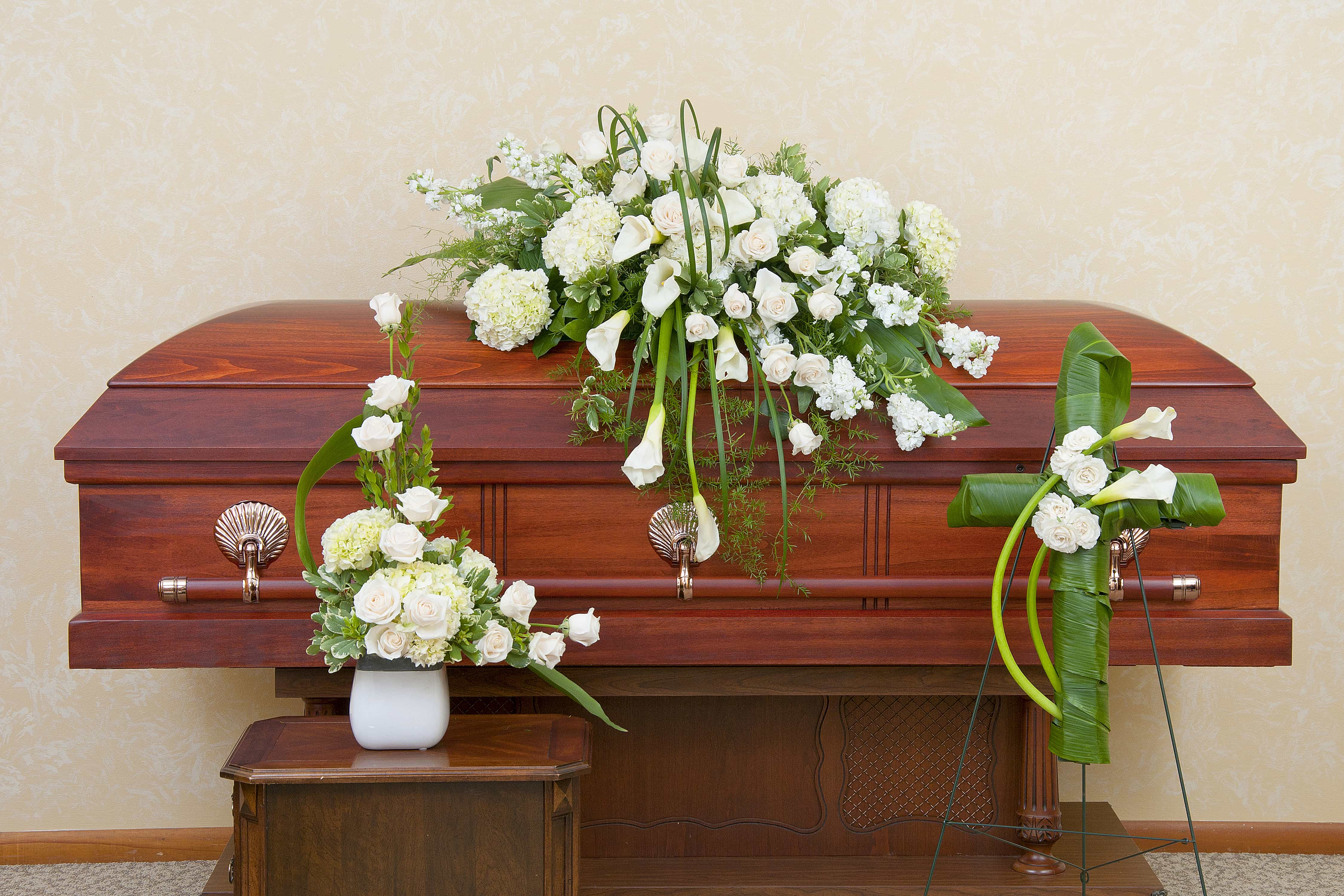 White floral funeral casket spray with matching arrangements beside a wooden casket