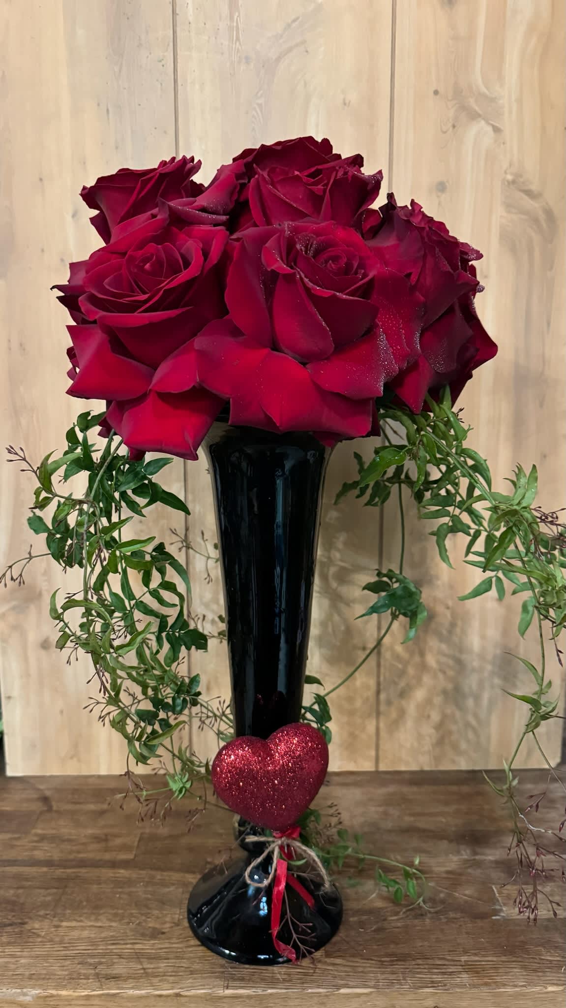 Red roses in a tall black vase with trailing greenery and a glittery heart