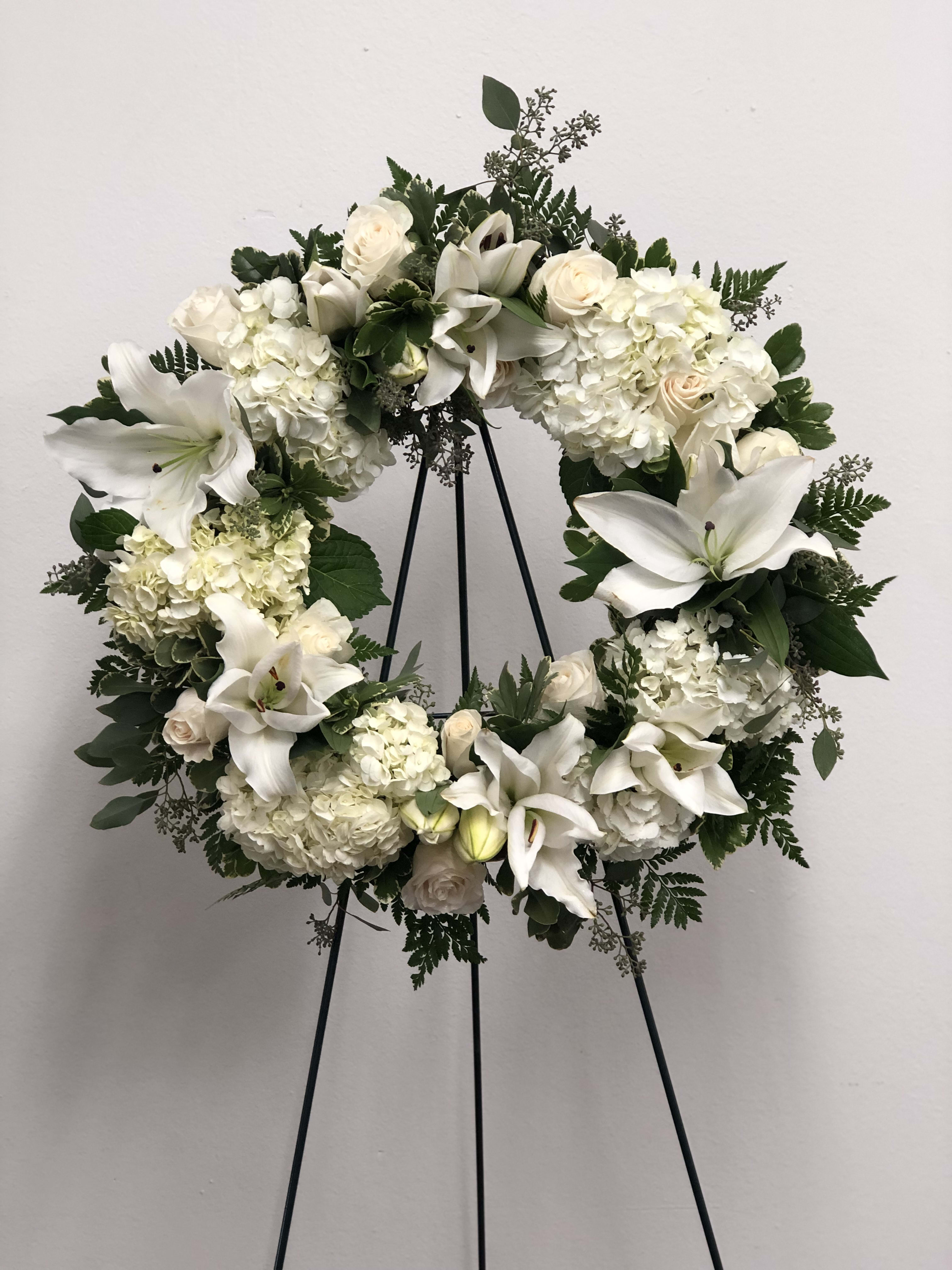 White floral wreath on a black stand with lilies, roses, and hydrangeas