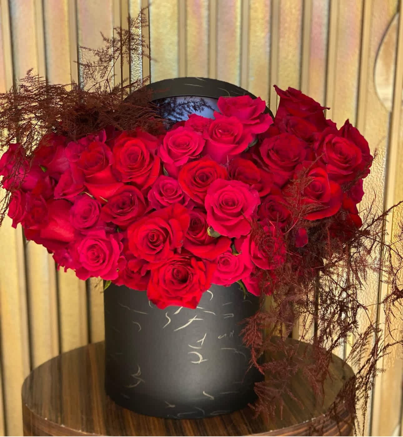 Red roses arranged in a black round box with dark feathery accents