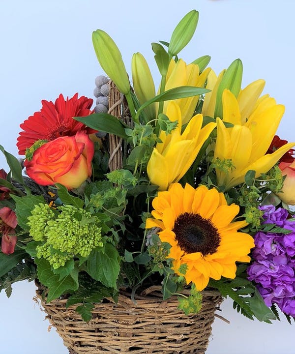 Mixed bouquet in a wicker basket with lilies, roses, sunflowers, and purple blooms