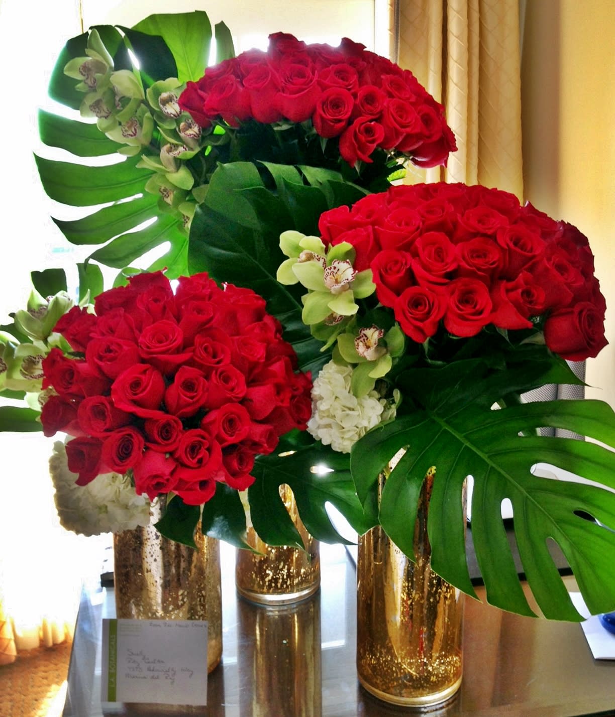 Red rose arrangements with orchids and large tropical leaves in gold vases