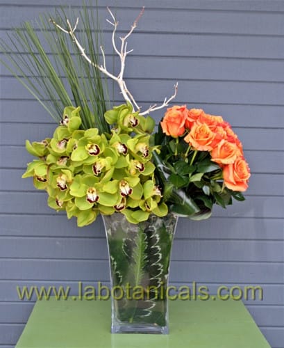 Orange roses and green orchids in a tall glass vase