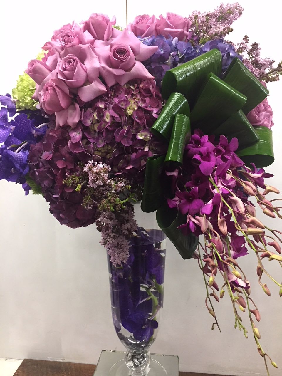 Purple roses and orchids arranged in a clear vase with a large green ribbon