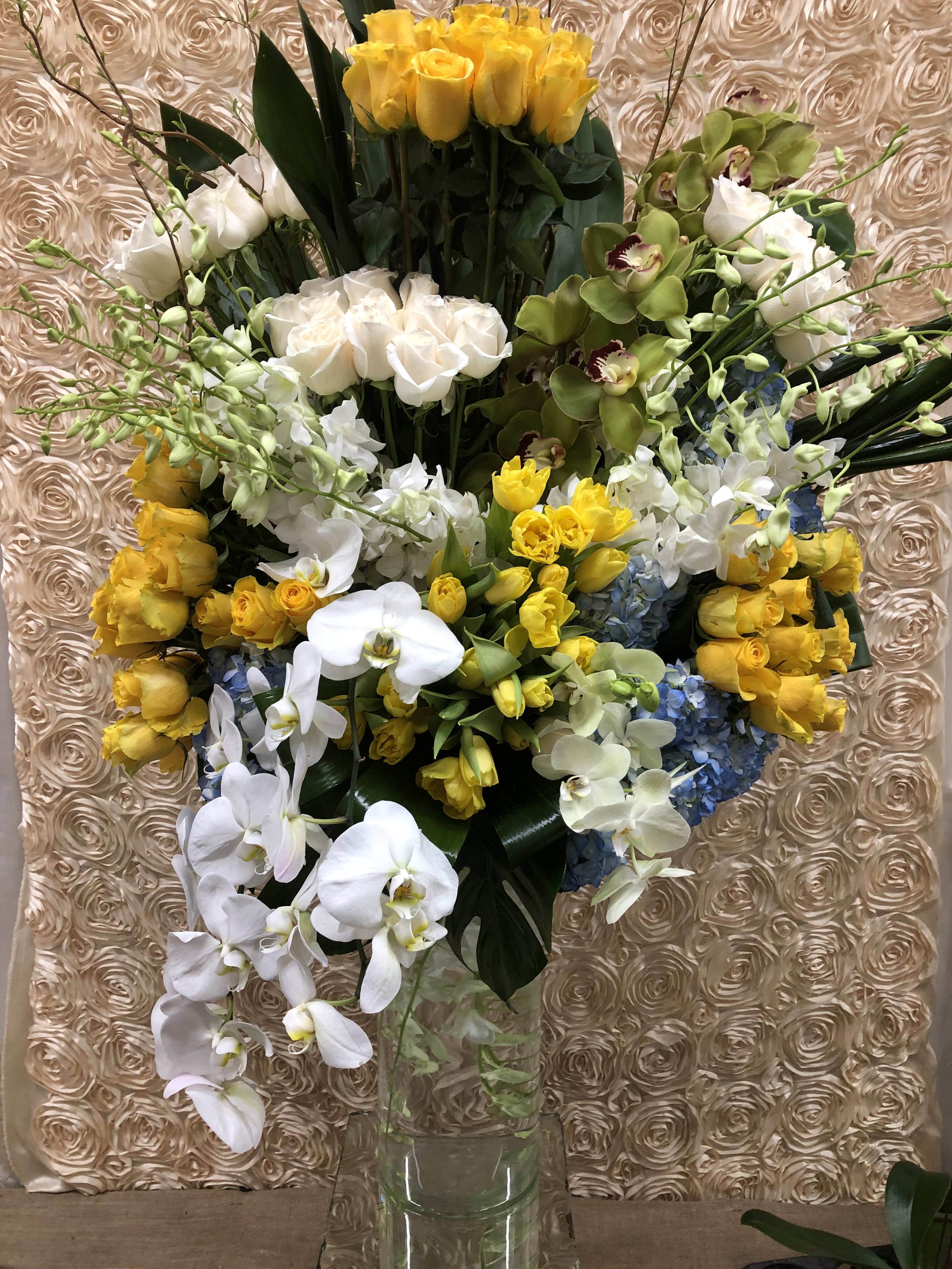 Tall bouquet of yellow roses, white orchids, and blue hydrangeas in a clear vase