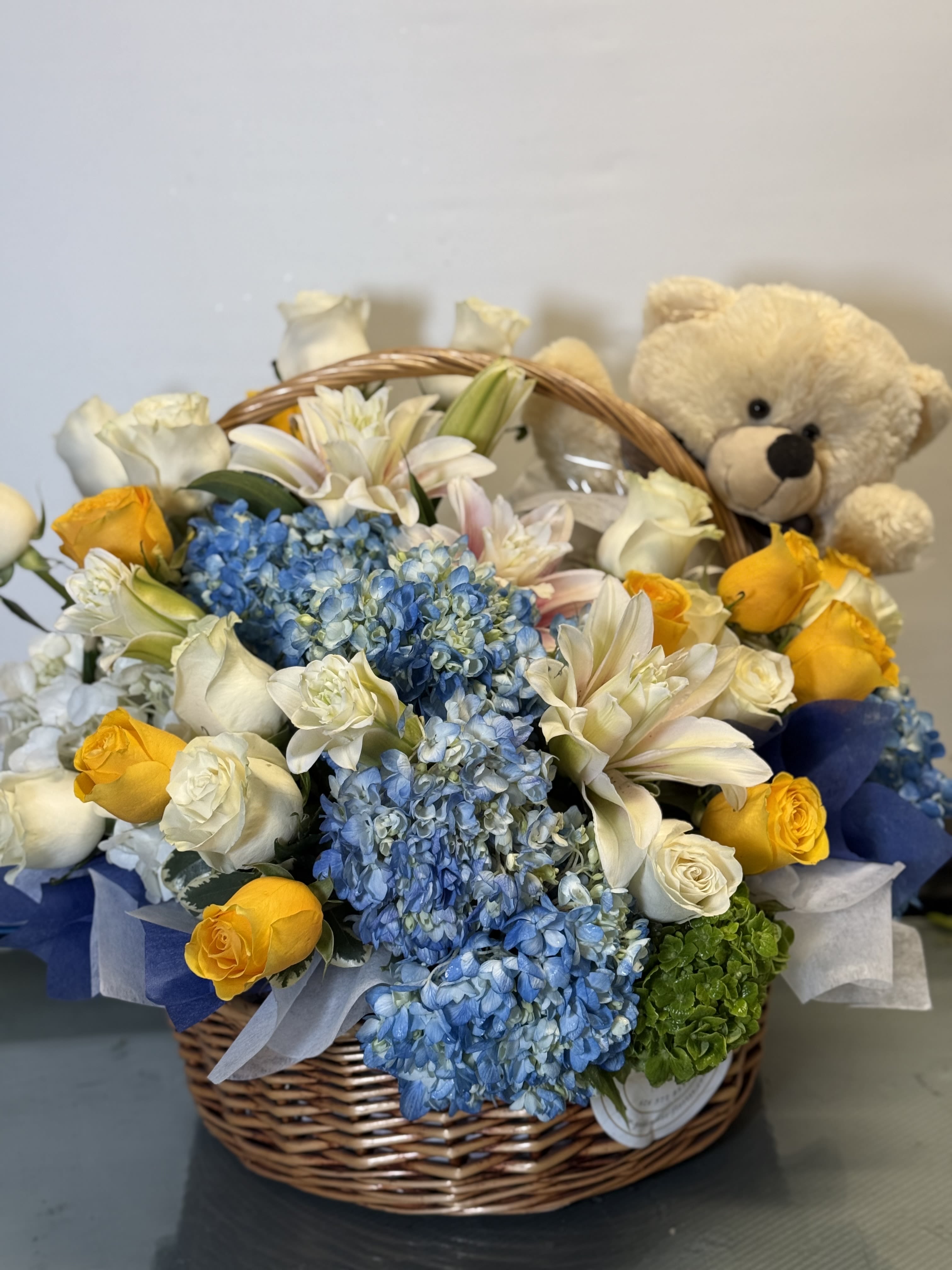 Basket of blue hydrangeas, white roses, yellow roses, and a teddy bear