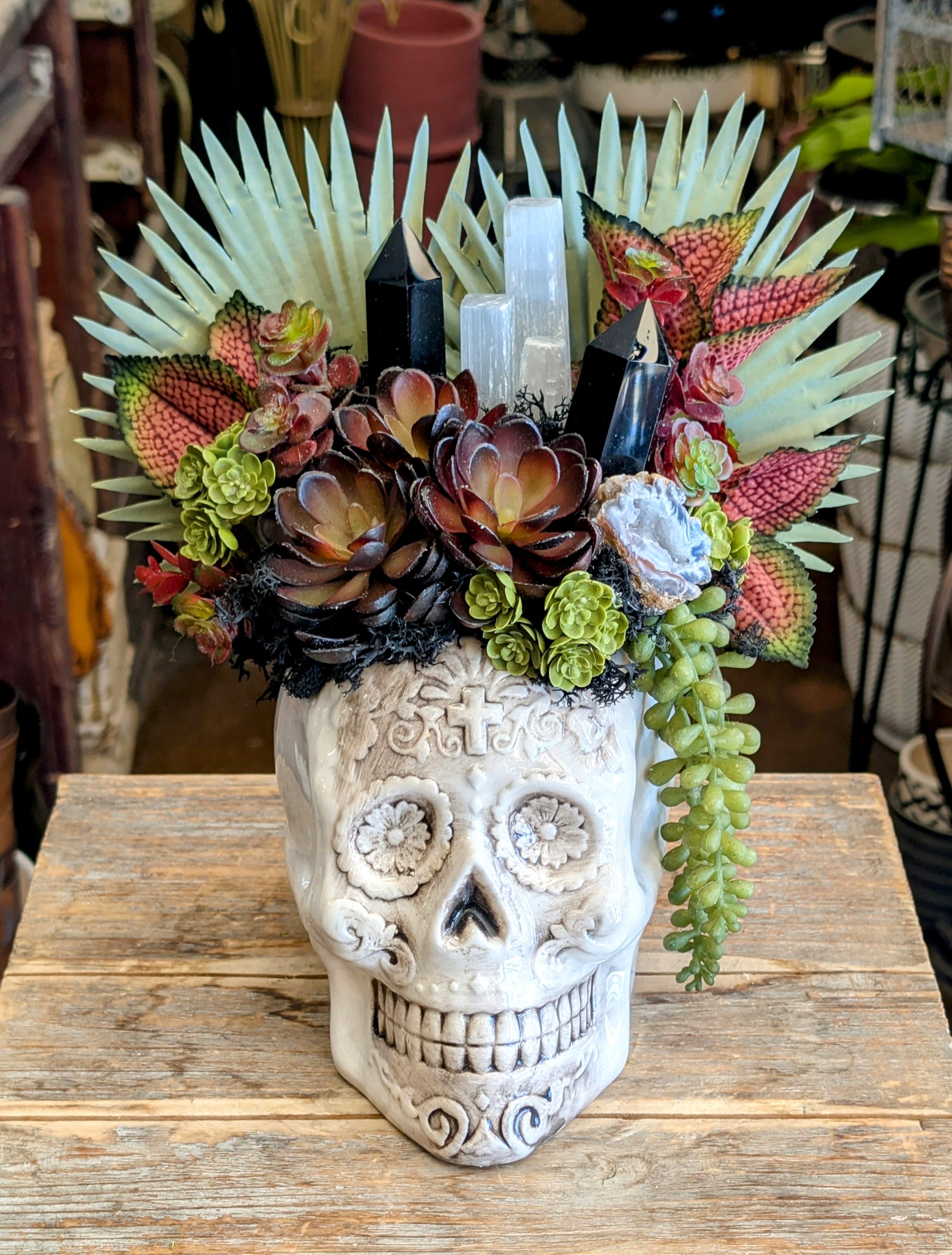 Eclipse- Faux Succulent And Crystal Arrangement - Eclipse – A Mystic Succulent Skull by Windmill Floral Expressions  Mysterious, bold, and steeped in celestial energy, Eclipse is a breathtaking statement piece from Windmill Floral Expressions in Las Vegas. This striking design rises from an intricately carved ceramic sugar skull planter, crowned with an extraordinary blend of faux succulents, preserved moss, exotic foliage, and shimmering crystals.  Deep burgundy echeveria and vibrant green rosettes spill over the skull’s crown, while trailing string‑of‑pearls cascades dramatically along the side. Black moss and textured leaves weave through spires of obsidian and selenite crystal towers, their light‑and‑shadow interplay evoking the mystery of an eclipse. A raw geode glimmers at the center, a hidden treasure radiating natural magic.  Perfect For: ️ A bold gothic centerpiece or altar focal point ️ Boho‑witchy home décor with striking artistry ️ An unforgettable gift for crystal lovers and those drawn to the mystical  With Eclipse, your space is transformed into a realm of fearless beauty, cosmic energy, and timeless intrigue—no watering required.   Handcrafted in Las Vegas, Eclipse is ready to bring its otherworldly charm into your home.  Same day delivery to Anthem,  Anthem Highlands, Inspirada, Mcdonald Ranch, Green Valley, Seven Hills, Roma Hills, Grand Legacy, The Fountains, Cadence, Lake Las Vegas and Ascaya.  