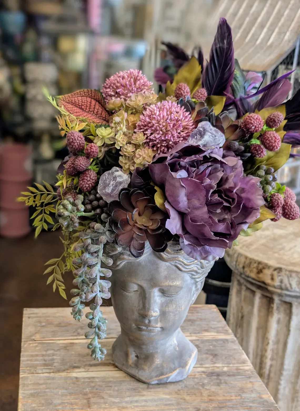 Viola Noir- Faux Botananical Arrangement - Unveil the dark romance of Viola Noir — a sculptural masterpiece of faux botanicals designed by Windmill Floral Expressions in Las Vegas, Nevada. This elegant arrangement blooms from a classical stone bust, crowned with deep violet peonies, succulents, berries, and lush botanical textures in tones of plum, sage, and wine. Subtle amethyst crystals and cascading greens add a mystical shimmer, creating a perfect fusion of bohemian artistry and modern sophistication.  Crafted entirely from luxury permanent botanicals, Viola Noir offers timeless beauty without maintenance — a showstopping centerpiece for Las Vegas homes, luxury offices, spas, or boutique interiors. Each detail reflects Windmill Floral Expressions’ signature style: imaginative, textural, and unmistakably artistic.