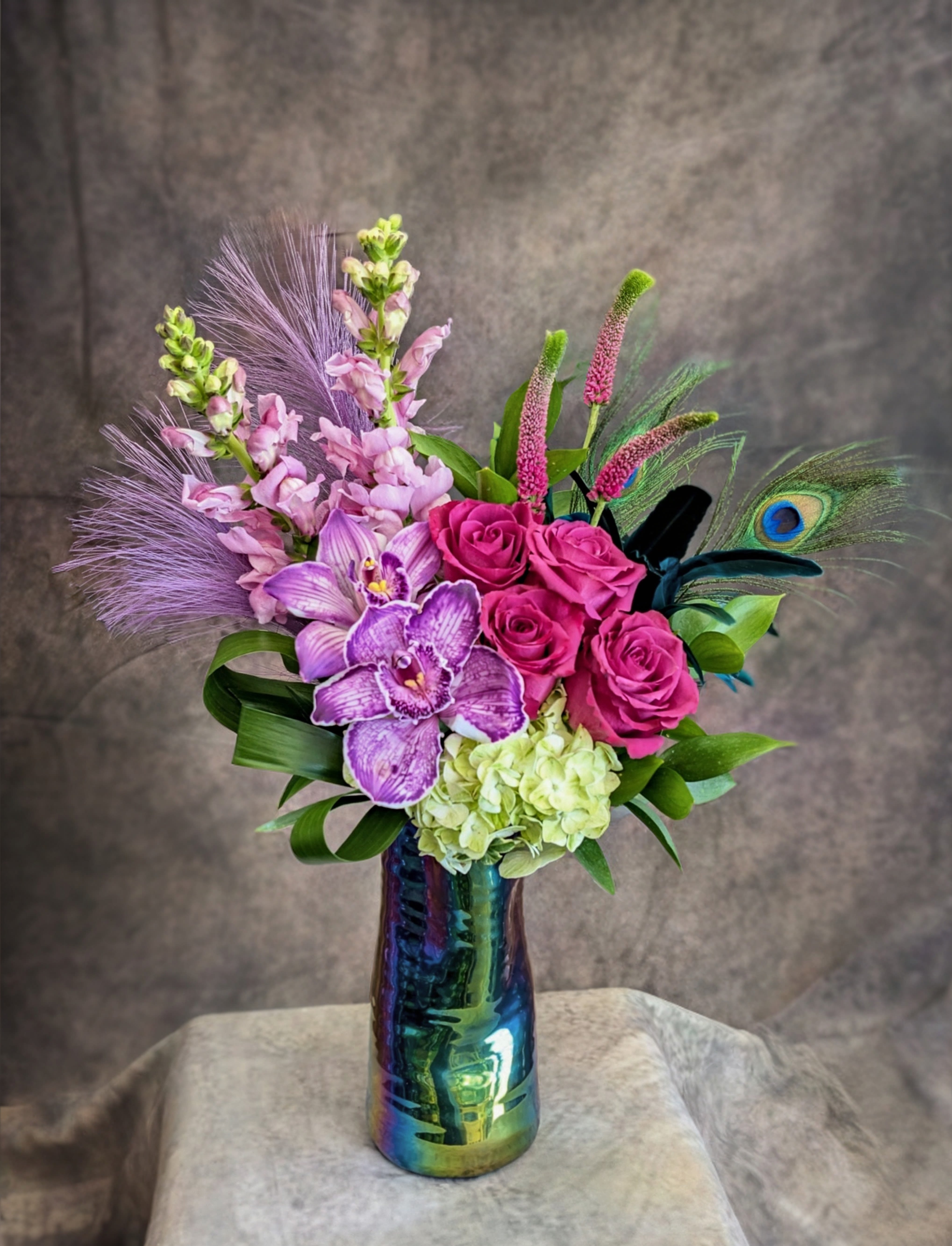 Zara - Bold, vibrant, and undeniably modern, Zara is a striking floral arrangement designed in rich jewel tones with an artful, exotic edge. Featuring premium Blueberry roses in deep plum hues, this design is accented with elegant purple cymbidium orchids, soft green hydrangea, and delicate snapdragons for a lush, dimensional composition. Textural touches of veronica, tropical greenery, and a dramatic peacock feather add movement, height, and a distinctive, high-end finish. Arranged in an artistic iridescent ceramic vase, Zara is perfect for birthdays, celebrations, or anyone who loves a bold, statement floral with a unique twist. Its vivid palette and sculptural design make it a standout choice for luxury flower delivery in Las Vegas. Handcrafted in Las Vegas by Windmill Floral Expressions, a locally owned and family-operated florist established in 1991, each arrangement is custom-made with the freshest flowers available—no premade designs, always one-of-a-kind.  Same-day flower delivery available in Las Vegas and Henderson, including Anthem, MacDonald Ranch, and surrounding areas.  Curbside pickup available—please call ahead to place your custom order. Perfect for: birthday flowers, modern floral arrangements, exotic-inspired designs, and luxury flower delivery in Las Vegas.