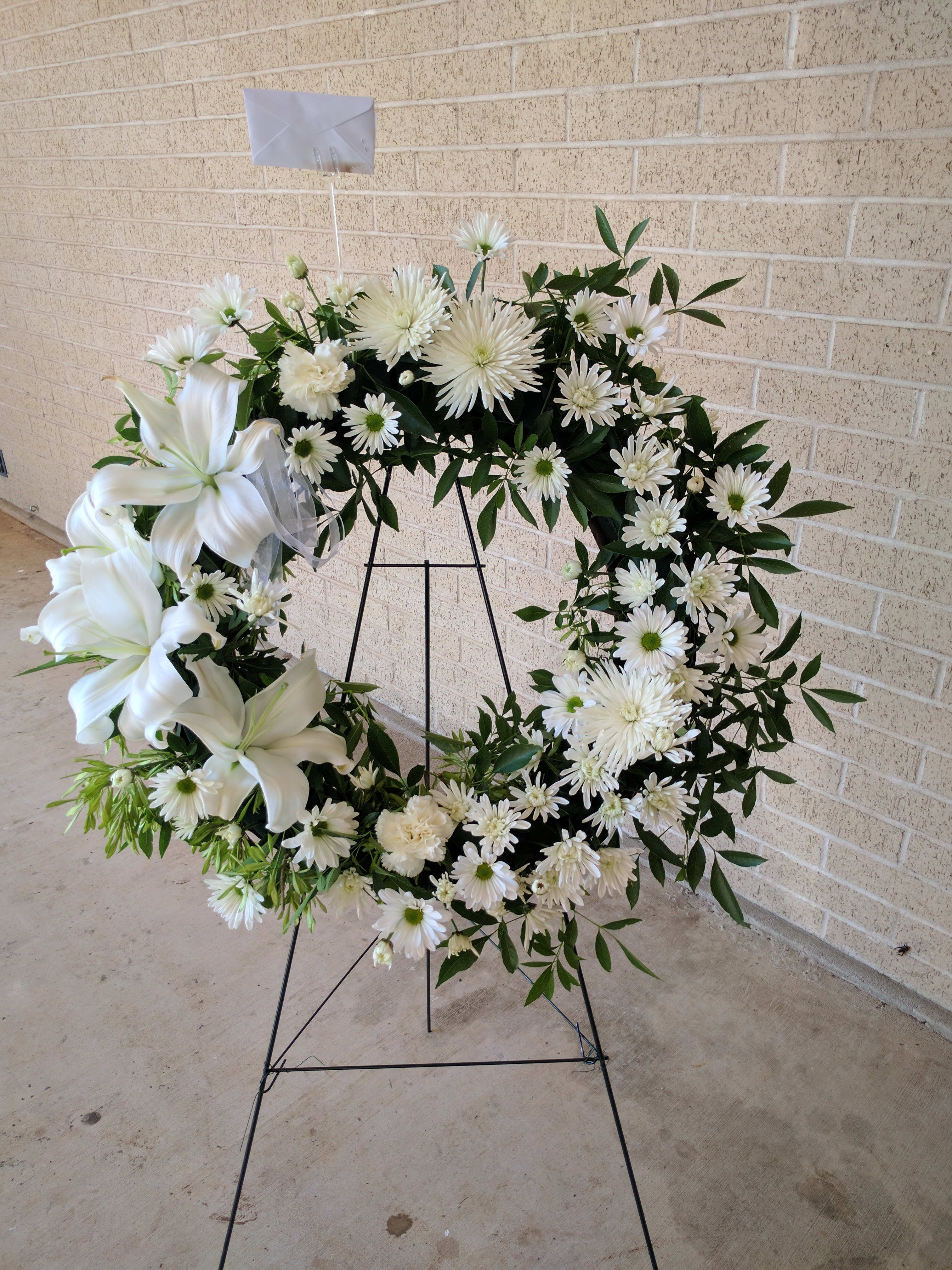 White floral wreath on a black stand with lilies and daisies